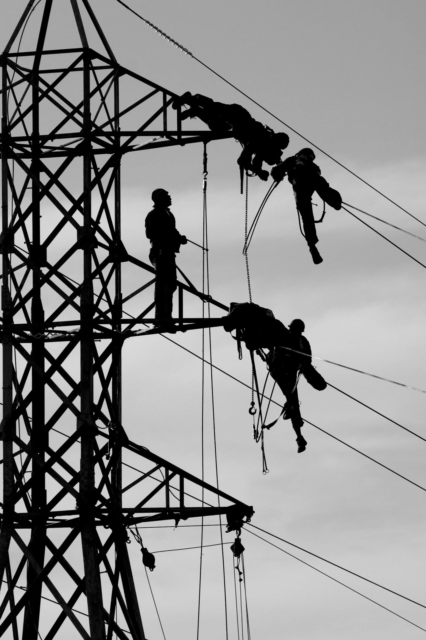 Silhouettes of workers performing maintenance or repairs on electrical power lines atop a transmission tower against a gray sky.