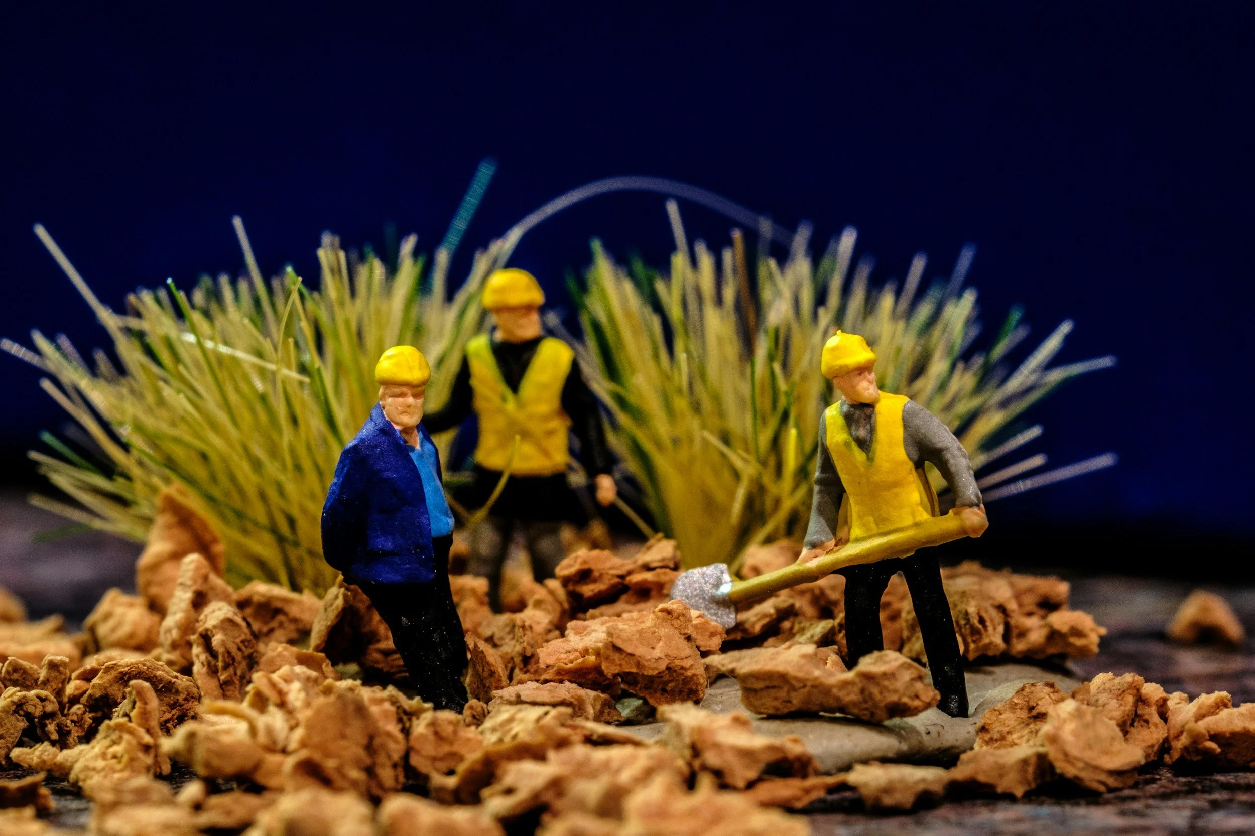 Miniature figurines depicting construction workers are placed on a rocky surface with artificial grass in the background, set against a dark blue backdrop.