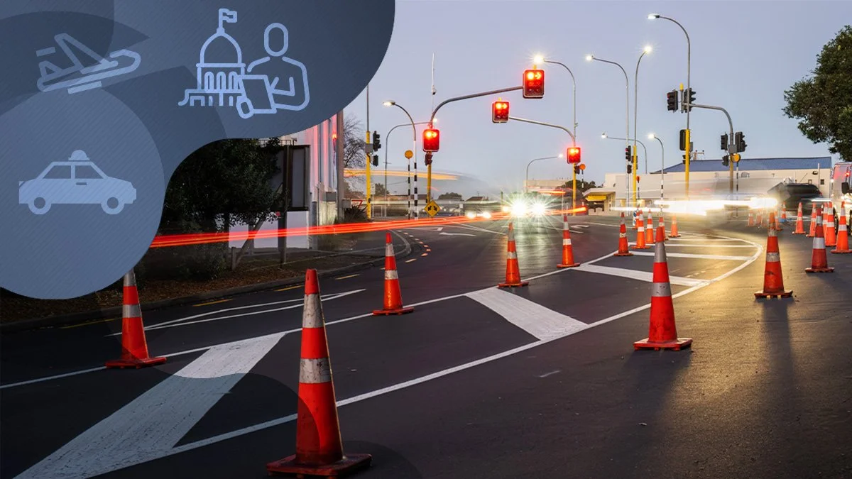 A city street intersection with orange traffic cones blocking the lanes, red traffic lights, and vehicle light trails indicating moving cars, with overcast evening sky.