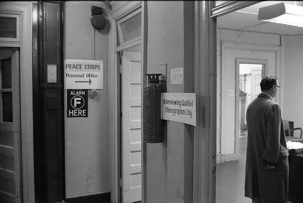 Interior of a building with signs indicating 'Peace Corps Personnel Office,' 'Alarm F Here,' and 'Interviewing Qualified Stenographers Only.' A man in a suit stands near a desk, and a fire extinguisher is mounted on the wall.
