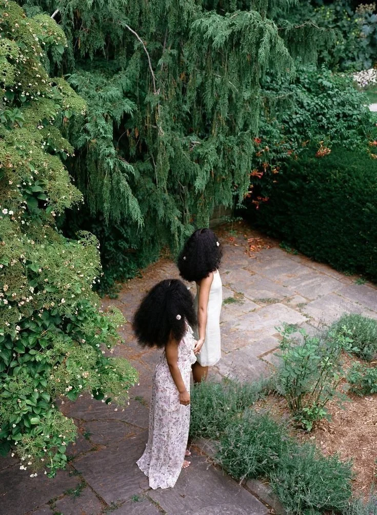 Two young girls with curly hair, one in a floral dress and the other in a white dress, stand hand-in-hand on a stone path in a lush garden surrounded by greenery and trees.
