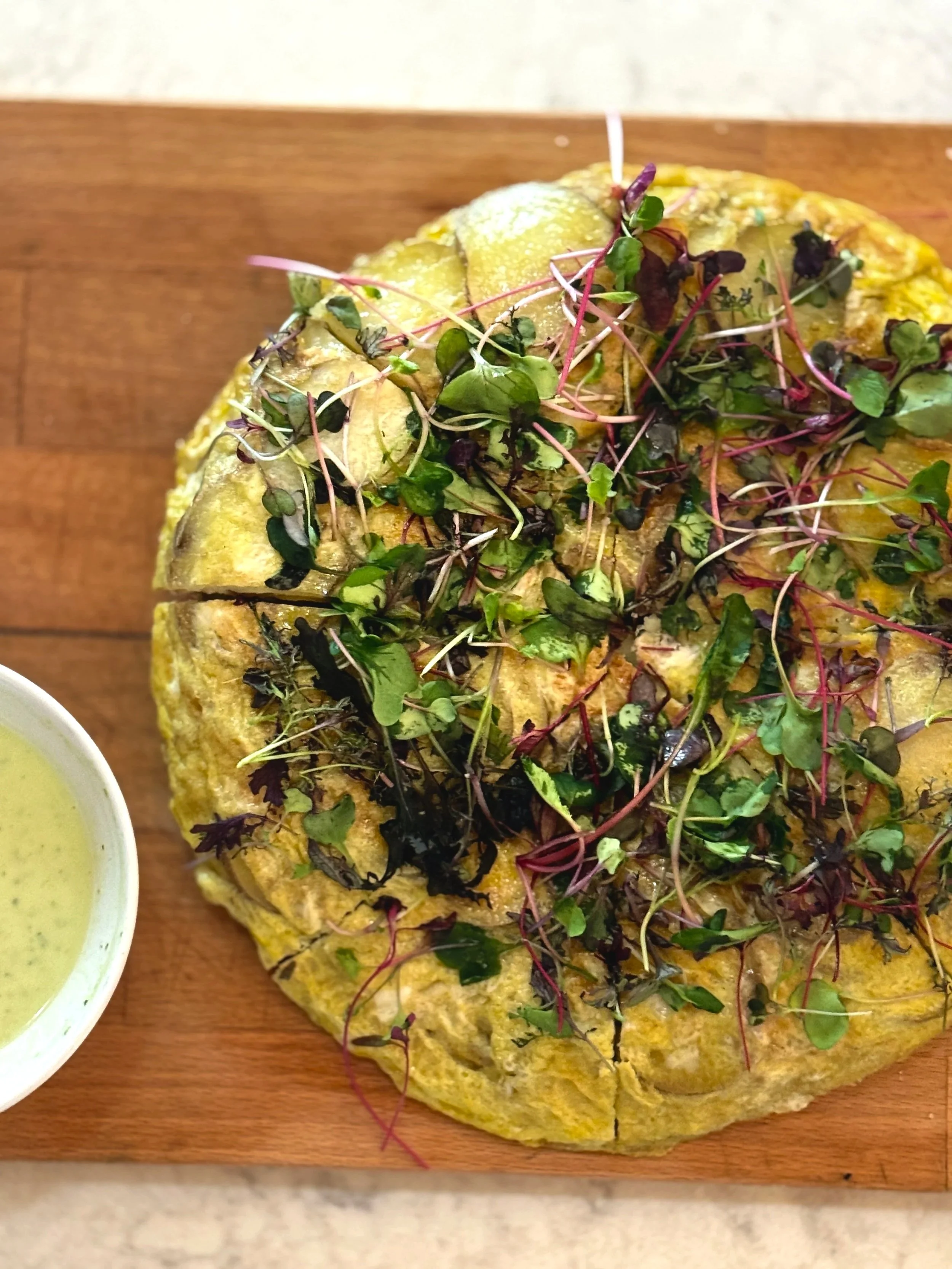 Baked dish topped with fresh microgreens and herbs, served on a wooden board with a small bowl of sauce
