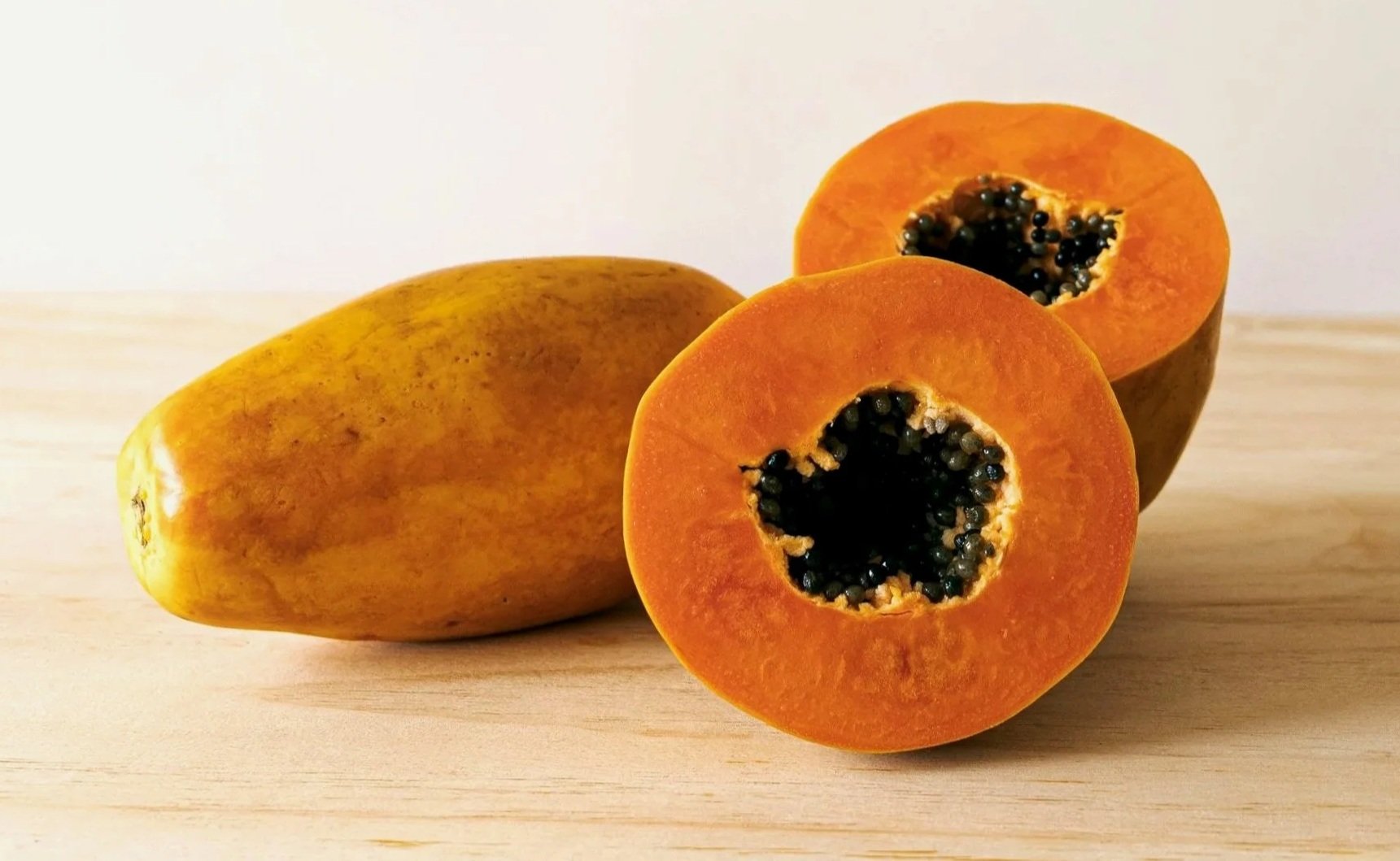 A whole papaya and a halved papaya displaying the orange flesh and black seeds inside on a wooden surface.