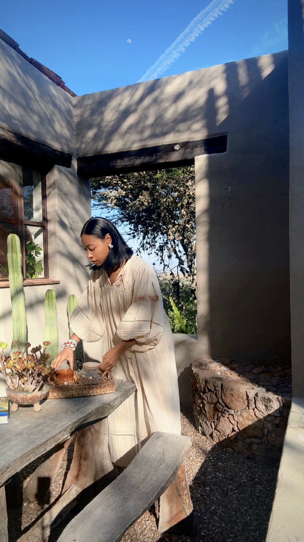 A woman in a beige dress arranging potted plants on a rustic wooden table outdoors. Sunlight casts shadows, with cacti and trees in the background and a clear blue sky overhead.