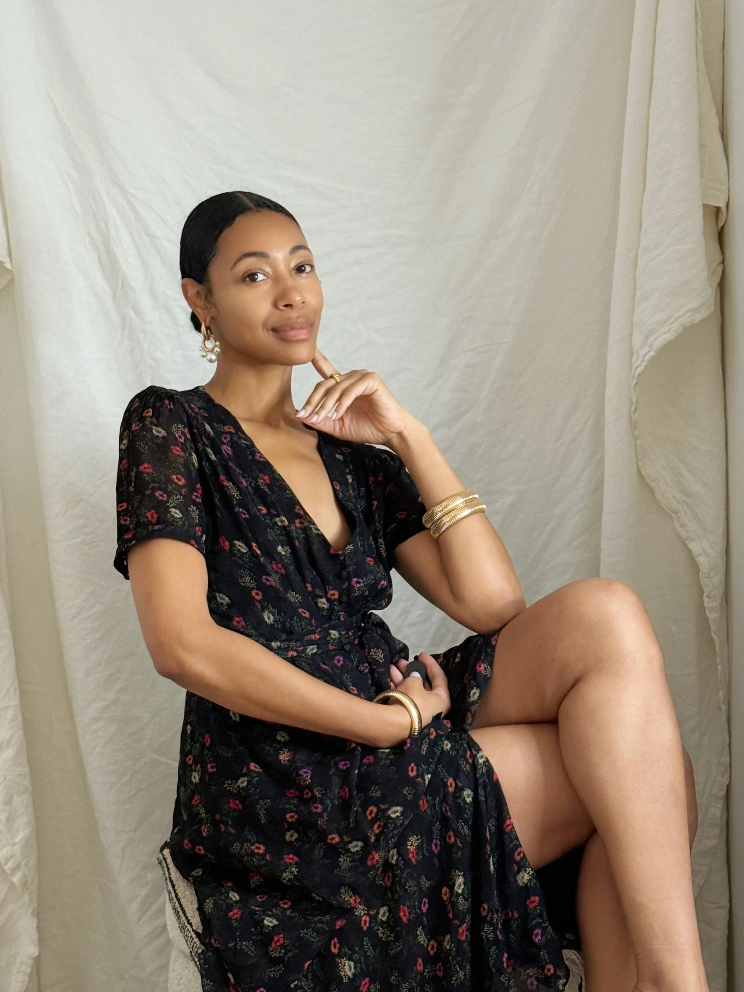 A woman with dark hair, wearing a black dress with floral patterns, sitting with one leg crossed over the other against a plain cream-colored backdrop, accessorized with gold earrings, bracelets, and rings.