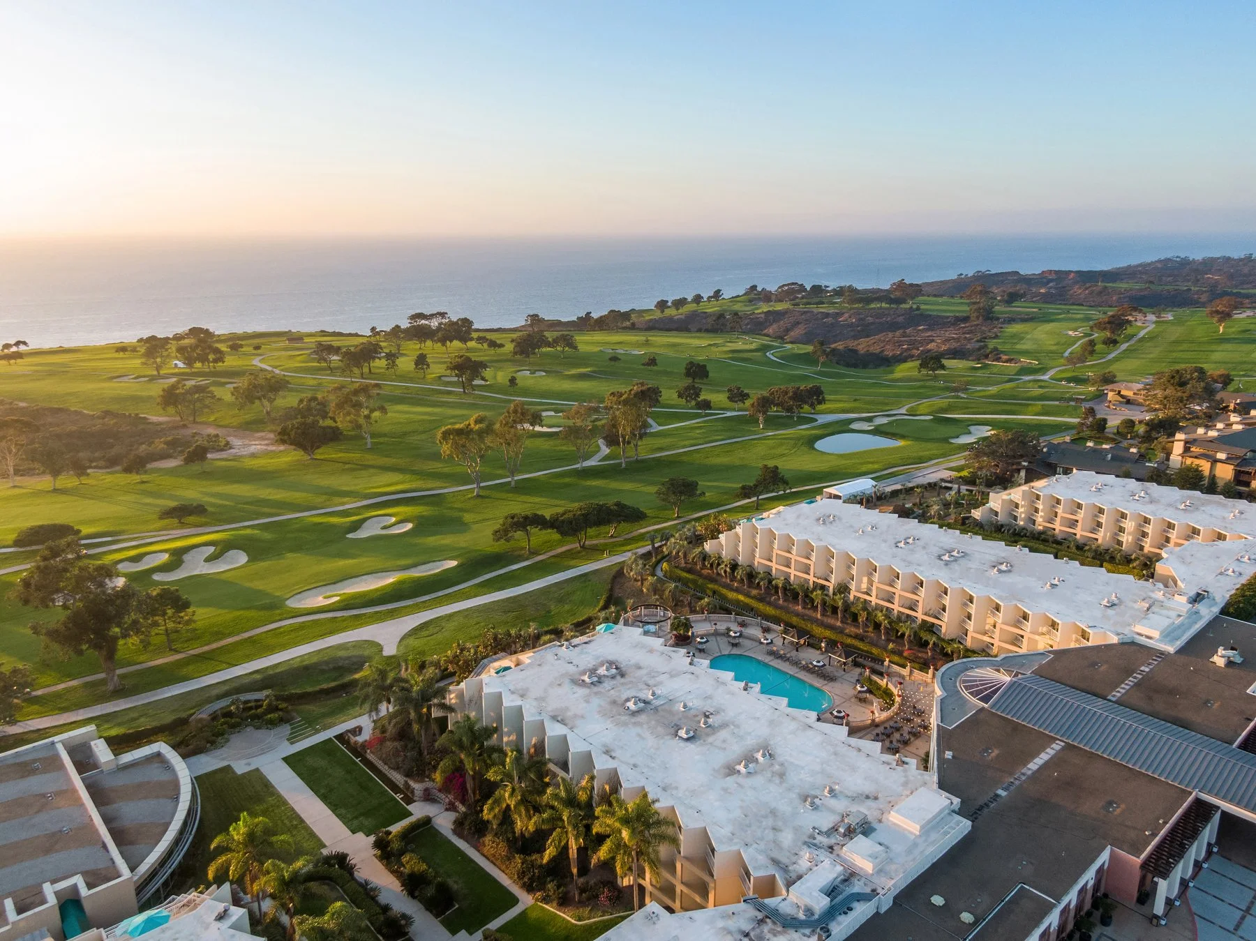 A scenic aerial view of a golf course near the coast with lush green fairways, sand bunkers, and trees, adjacent to a residential complex with swimming pools and white rooftops, during sunset.