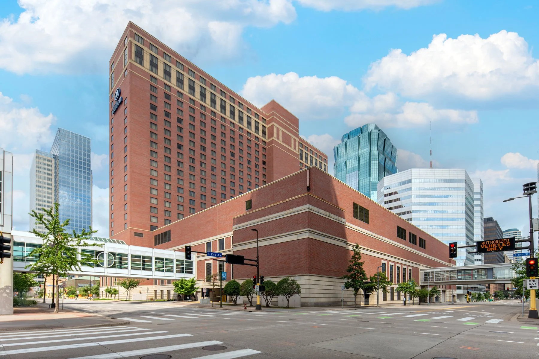 City street view featuring a large brick hotel building with multiple floors, surrounded by modern glass skyscrapers under a blue sky with scattered clouds.