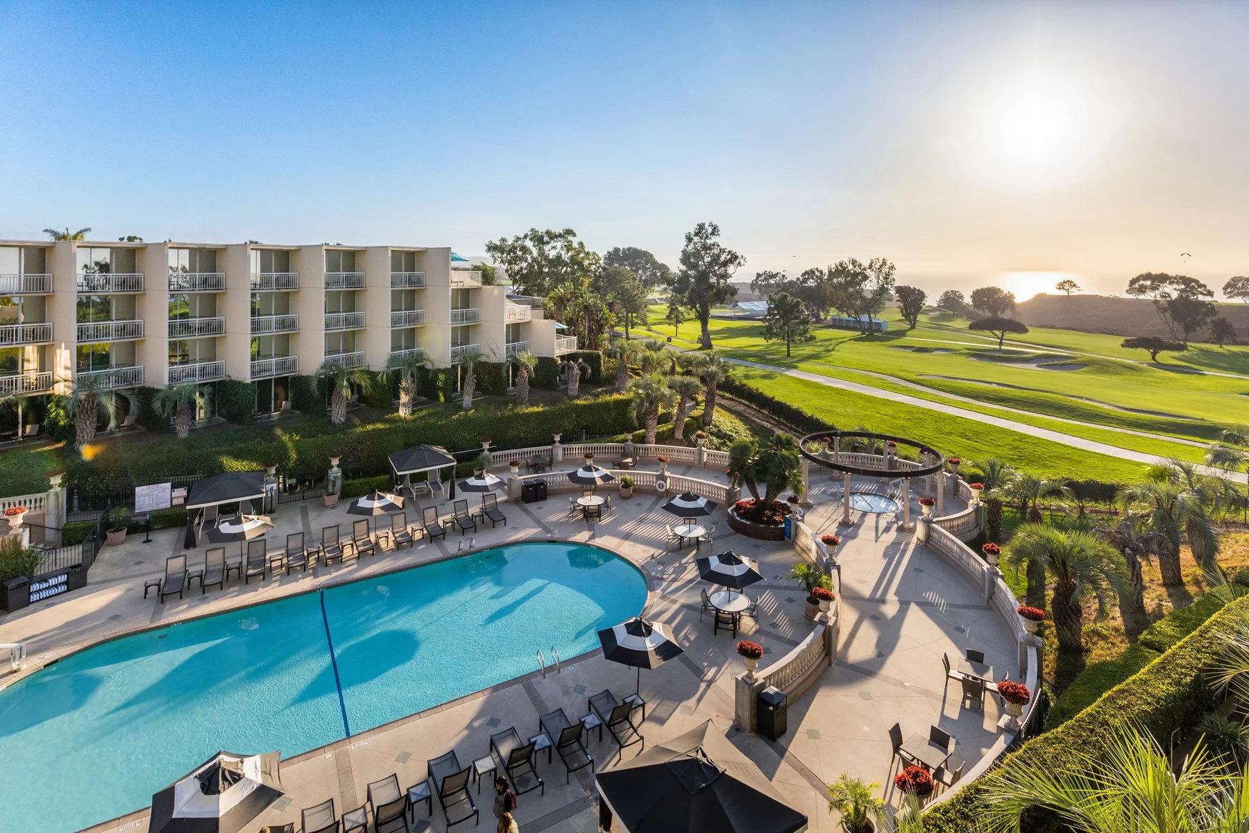 Sunset view of a resort with swimming pool, lounge chairs, umbrellas, and a golf course in the background.