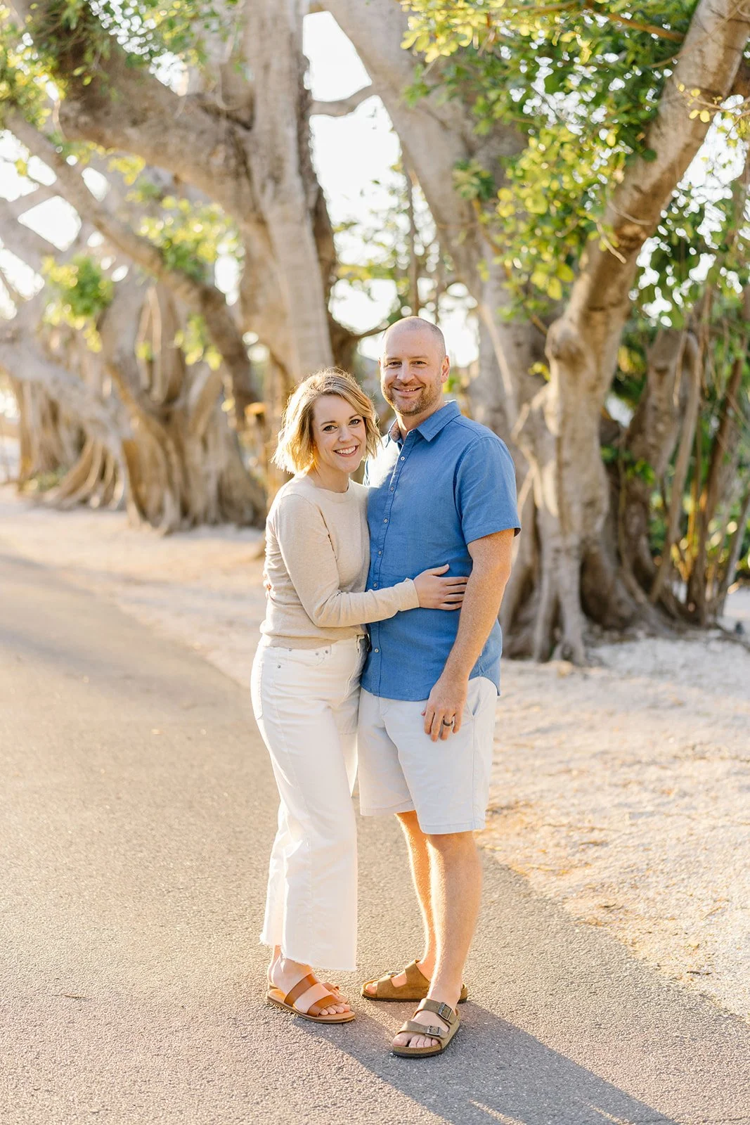 A smiling young woman and a man stand close together on a paved path, surrounded by large trees with twisted trunks, during the golden hour of sunset.