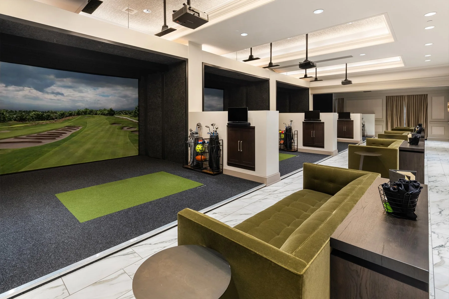 Indoor golf simulation room with multiple simulation bays, golf clubs, TVs, seating area, and a golf course projected on the screen.