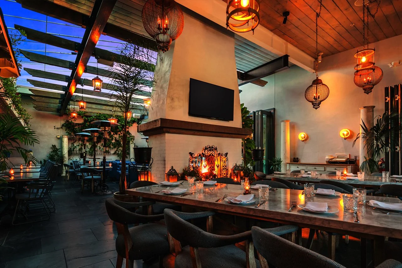 Interior of a restaurant with a cozy fireplace, wooden ceiling, warm lighting from hanging lanterns and sconces, tables set with napkins and glasses, and plants for decoration.