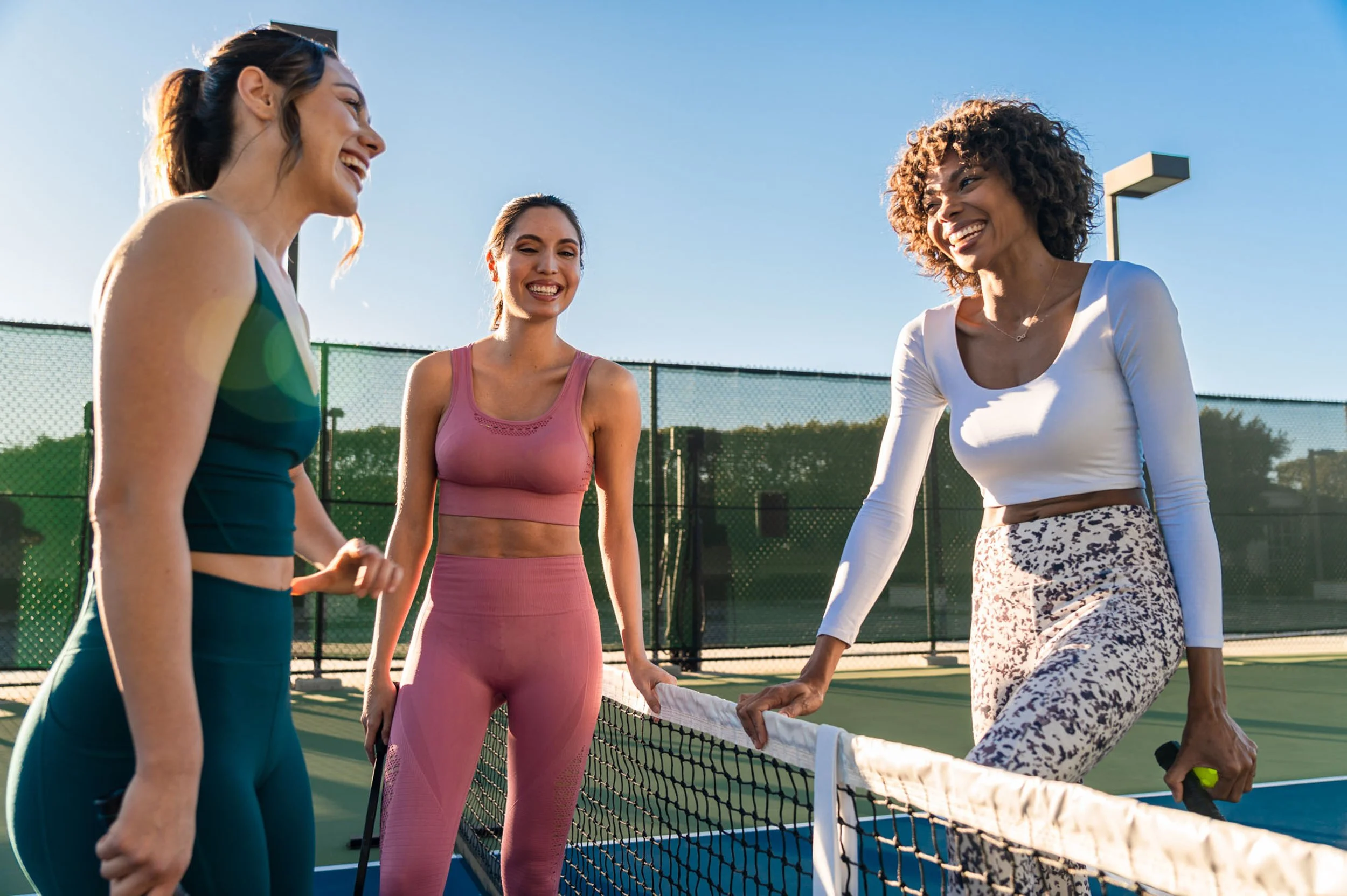 Three women standing on a tennis court, chatting and smiling during daytime, with tennis rackets in their hands.