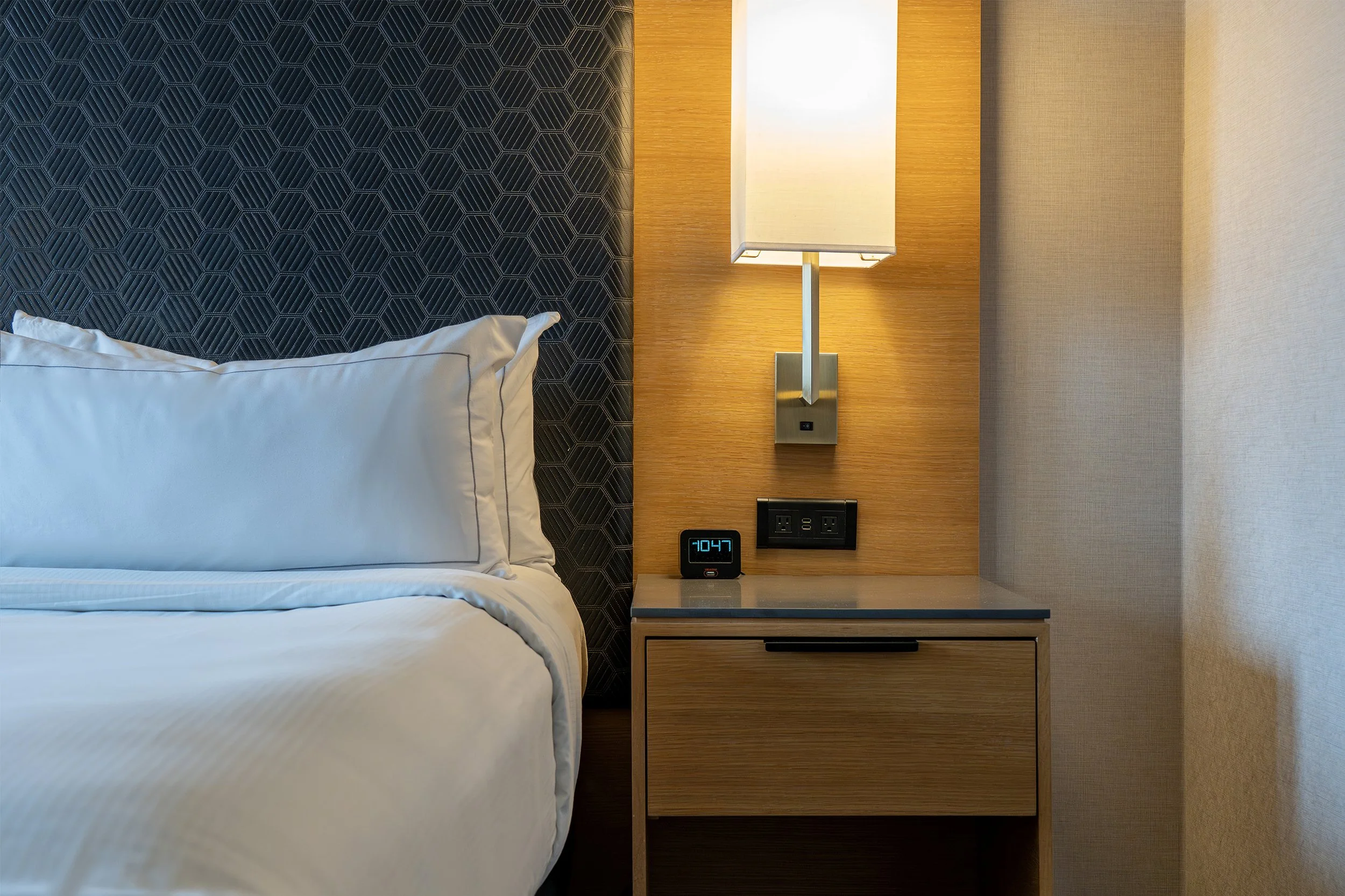A bedroom with a white bed and pillow, a gold-colored wall with a wall lamp, a digital alarm clock showing 10:47, and a nightstand underneath the lamp.
