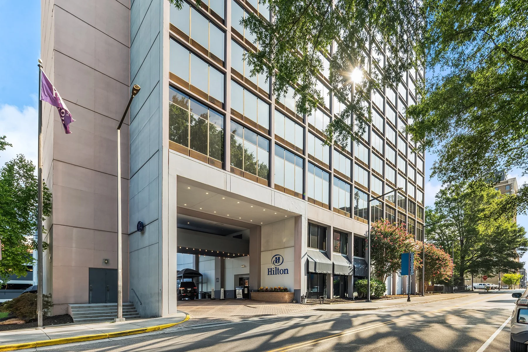 Exterior view of a Hilton hotel building on a sunny day with trees and cars parked on the street.