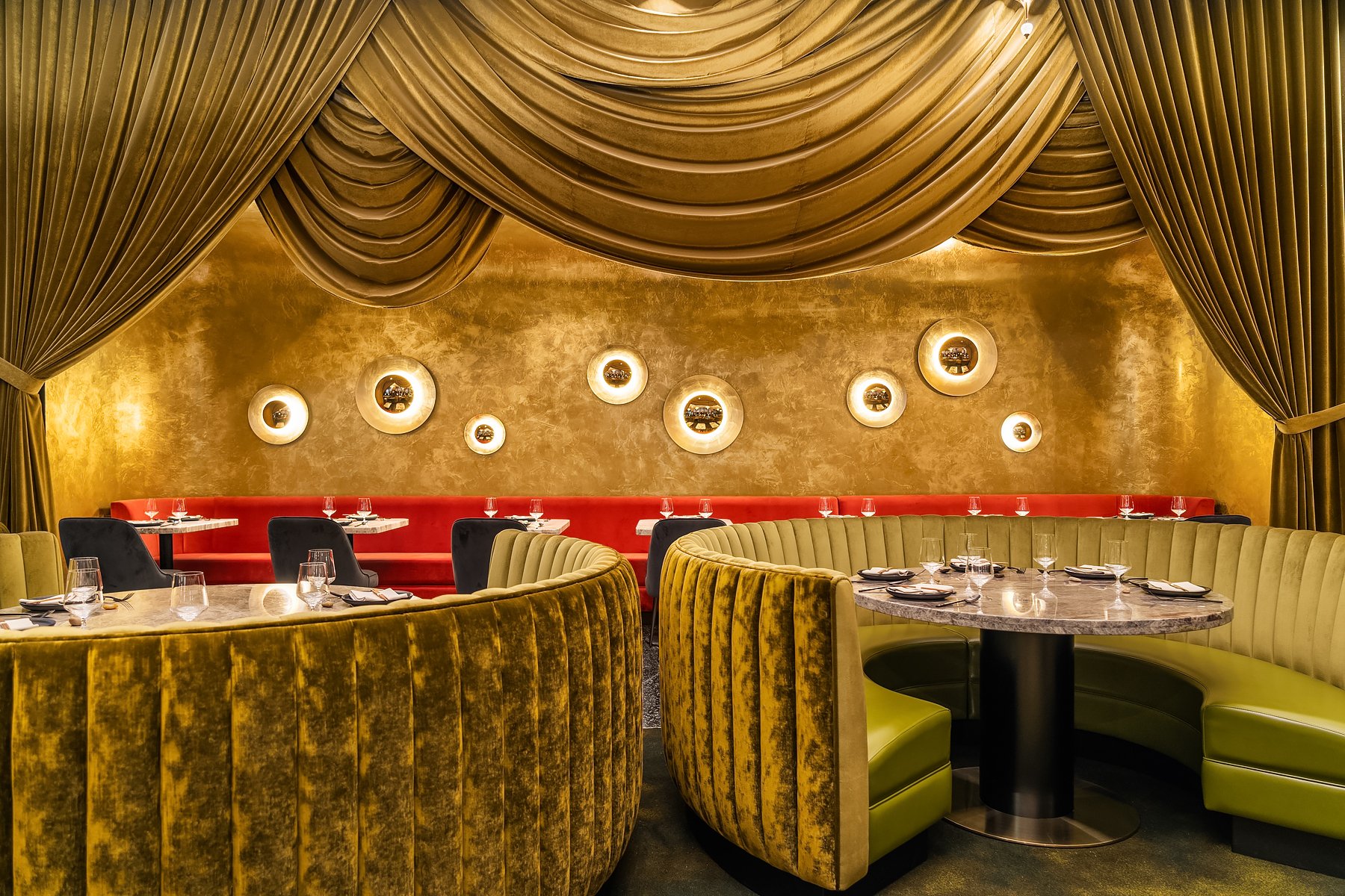 Elegant restaurant interior with gold drapery, gold textured wall, and circular wall lights framing pictures. The foreground features curved booths with yellow and green upholstery surrounding marble-top tables set with glassware and plates.