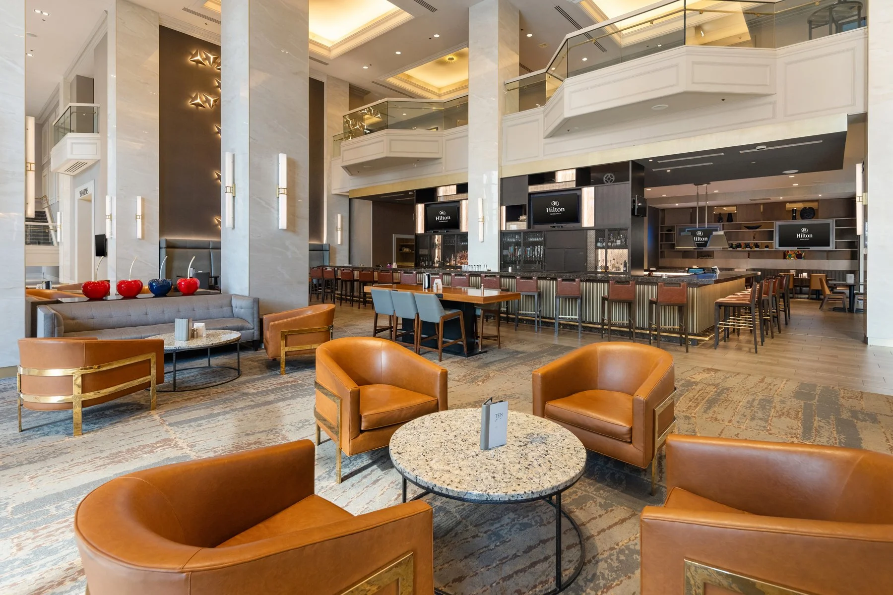 Luxury hotel lobby with comfortable chairs and couches, a bar area with barstools, multiple television screens, modern lighting, and elegant decor.