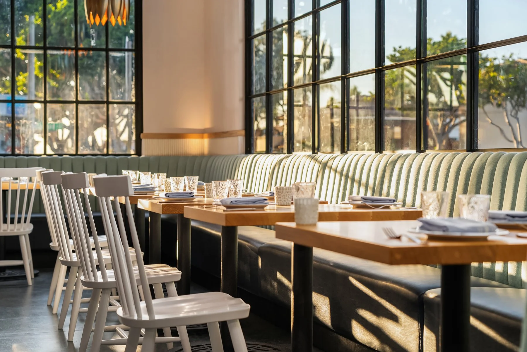 Restaurant with wooden tables, white chairs, a green upholstered banquette, and large windows allowing natural light inside.