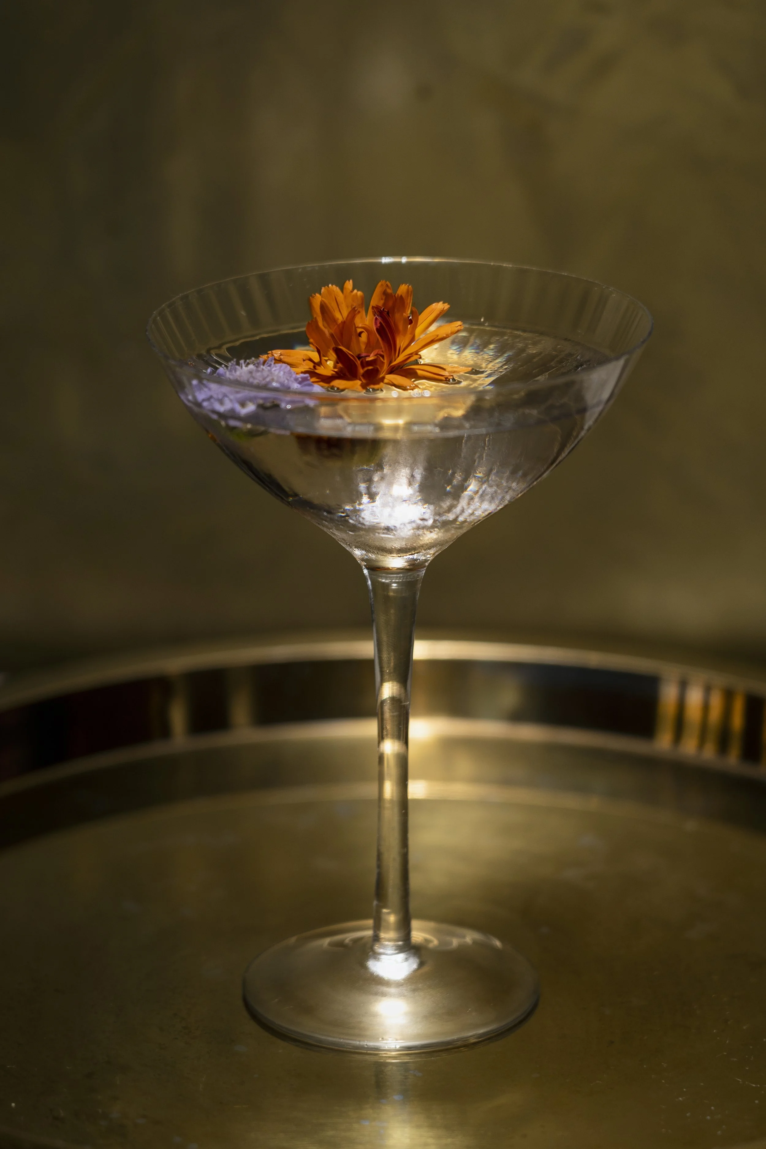 A cocktail garnished with an orange flower floating on top, placed on a reflective surface.