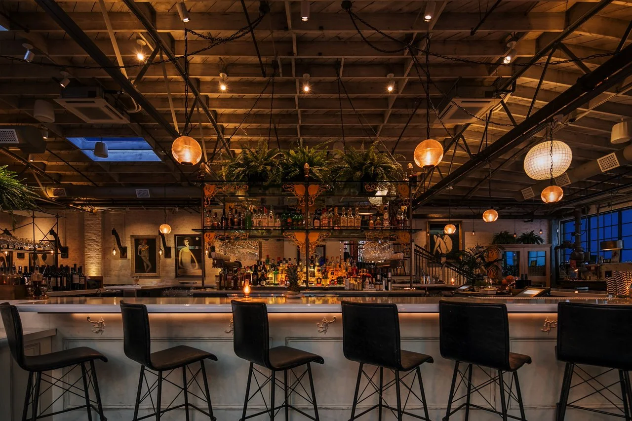 Interior of a dimly lit bar with a white marble counter, black bar stools, and warm hanging lights, decorated with potted plants and artwork