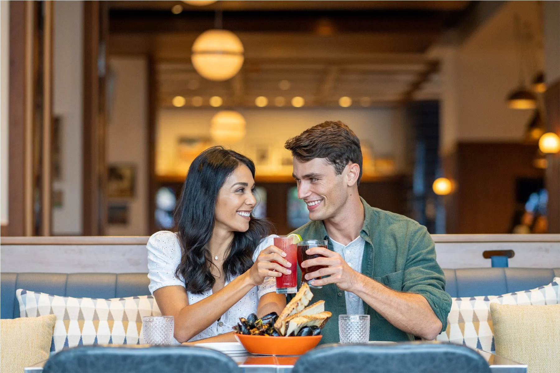 A smiling woman and man clinking glasses of cocktails in a restaurant, with a bowl of chips and dip on the table.