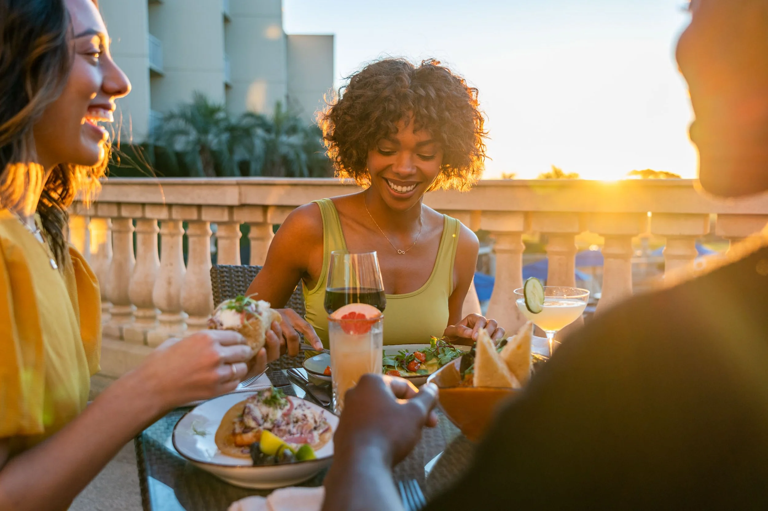 A group of friends enjoying a meal on an outdoor patio at sunset, with food and drinks on the table.