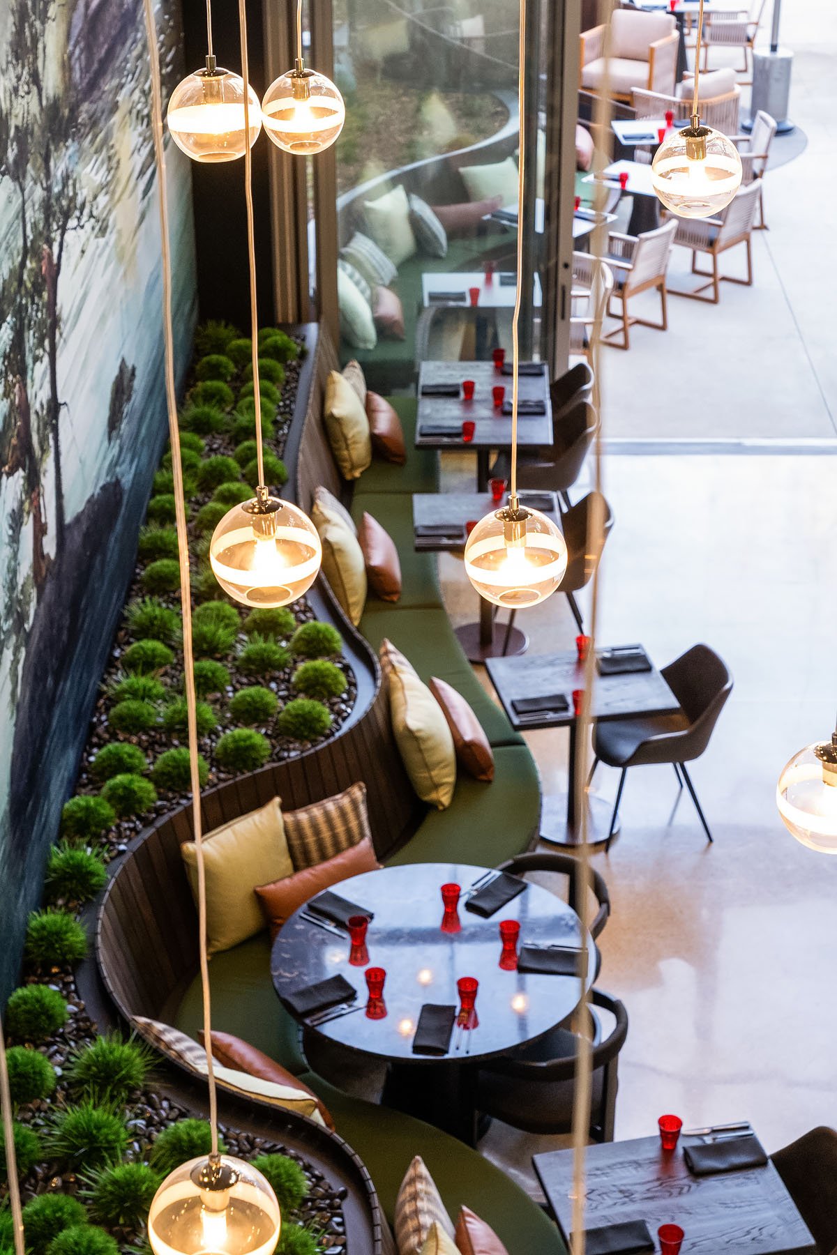 Interior of a restaurant with round tables set with black napkins, red glasses, and utensils. There's a curved green bench with various cushions, decorative planters with green plants, and modern hanging light fixtures.