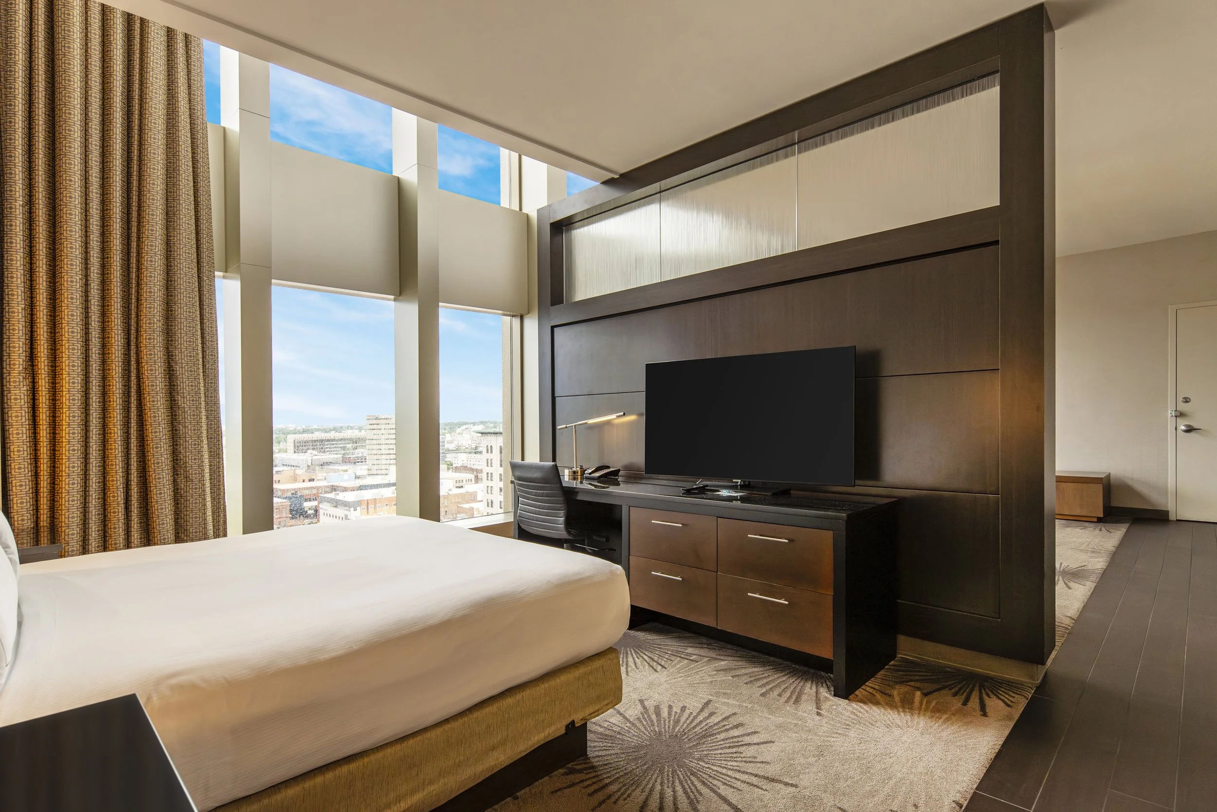 Hotel room with a large window, a bed with white bedding, a dark wooden dresser with a flat-screen TV, a desk with a lamp, and city skyline view