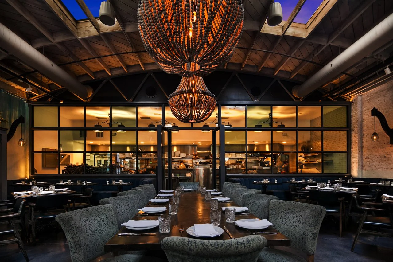 Elegant restaurant interior with a long dining table set with white plates, napkins, and glasses, surrounded by upholstered chairs. Two large chandeliers hang from a wooden ceiling, with a view into the open kitchen through a glass partition.