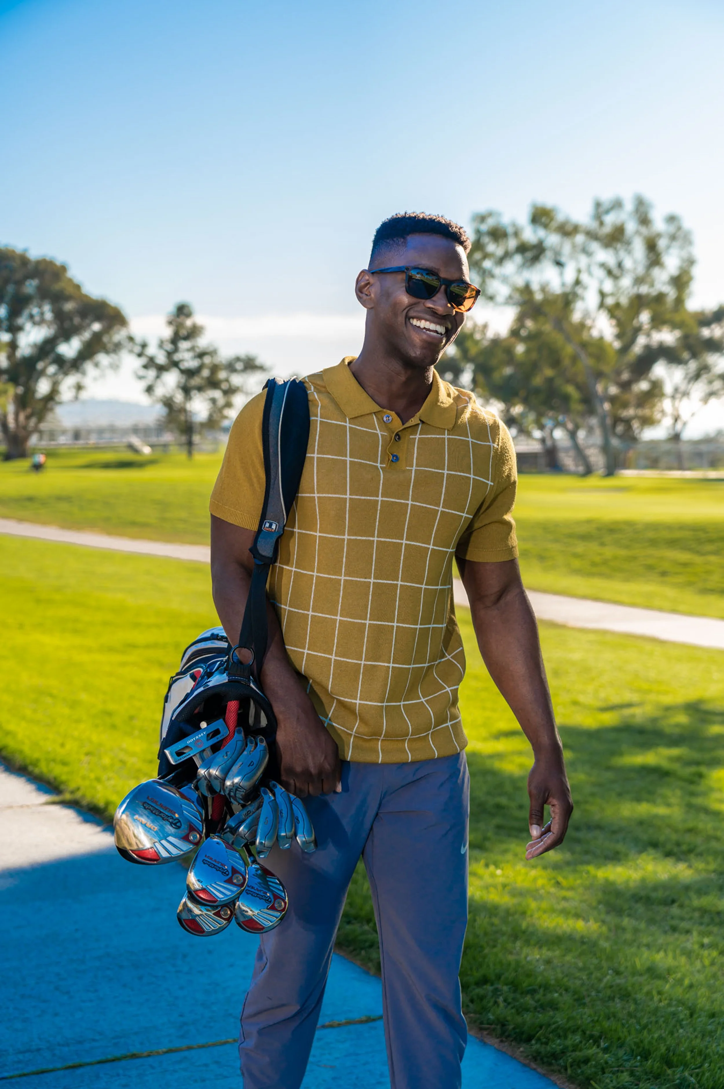 A young man wearing sunglasses, a yellow checkered polo shirt, and gray pants, holding a golf bag with golf clubs, standing on a golf course with trees and a blue sky.