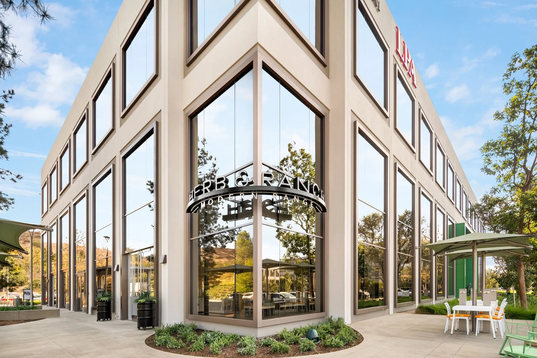 Exterior view of a modern commercial building with large glass windows, retail stores, outdoor seating, and trees, under a blue sky with some clouds.