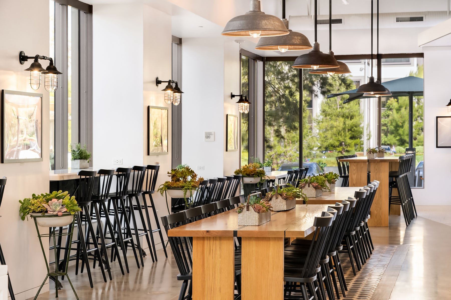 Interior of a modern food hall with a long wooden communal table decorated with plants, black chairs, high stools along the windows, and industrial pendant lighting. Large windows reveal a view of trees and outdoor seating.