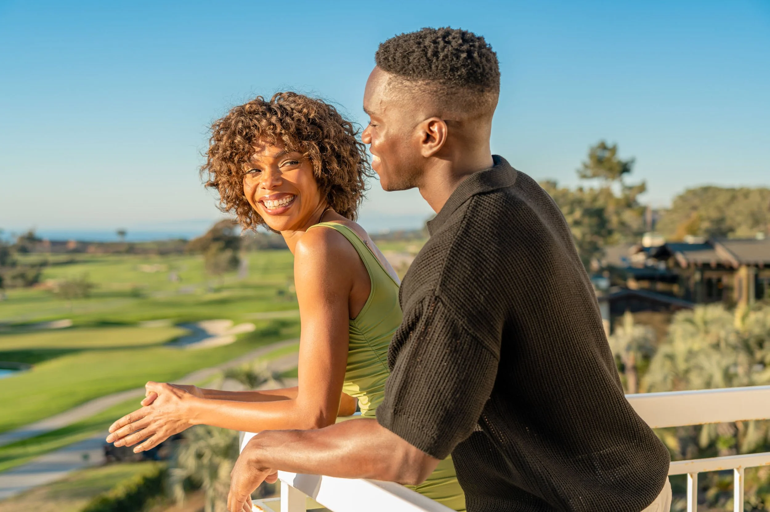 A man and woman smiling and talking outdoor on a sunny day with a scenic landscape of trees, grass, and a body of water in the background.