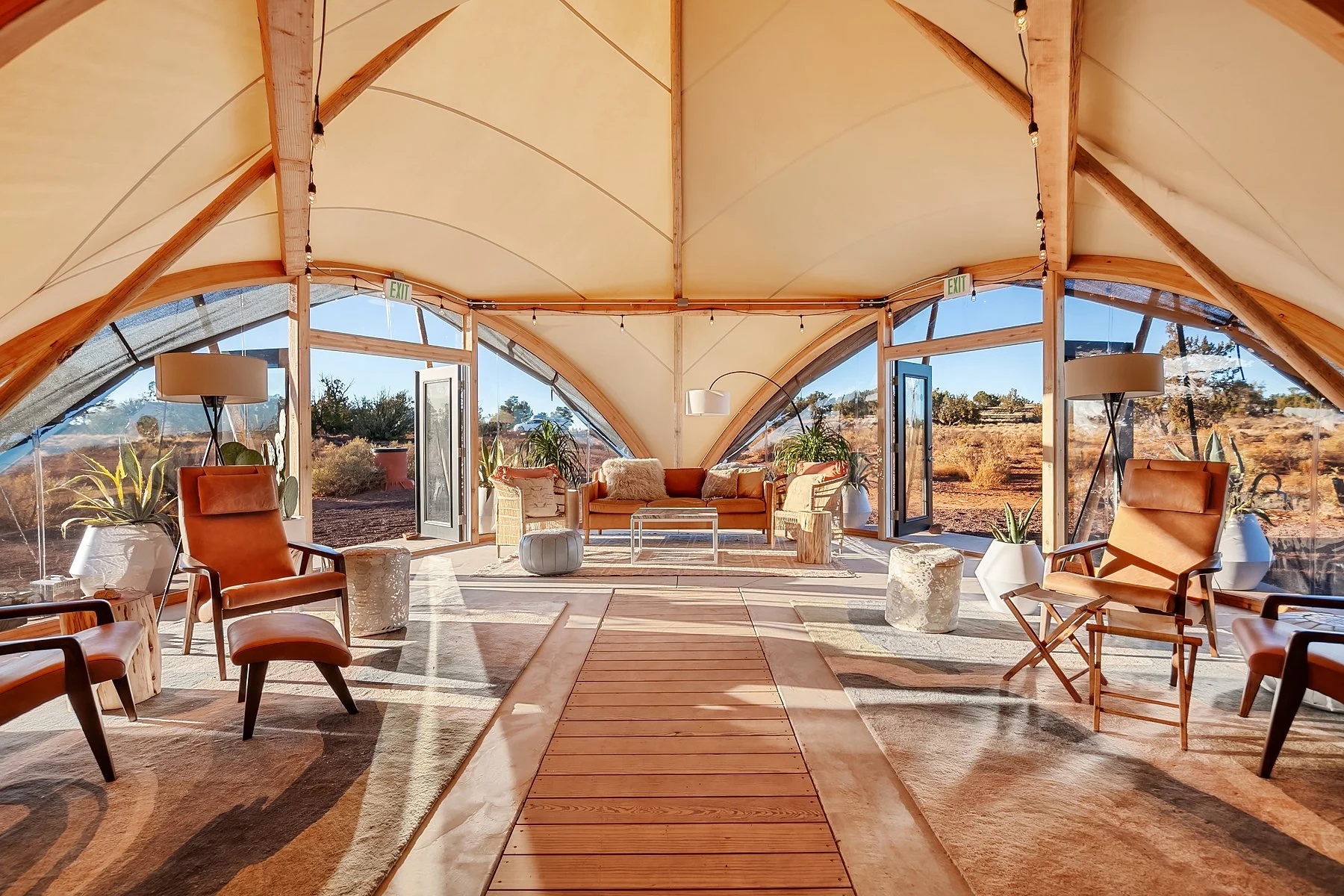 Interior of a modern, desert-themed living space with large, curved glass windows, wooden beams, leather chairs, a sofa, and outdoor view of desert landscape.