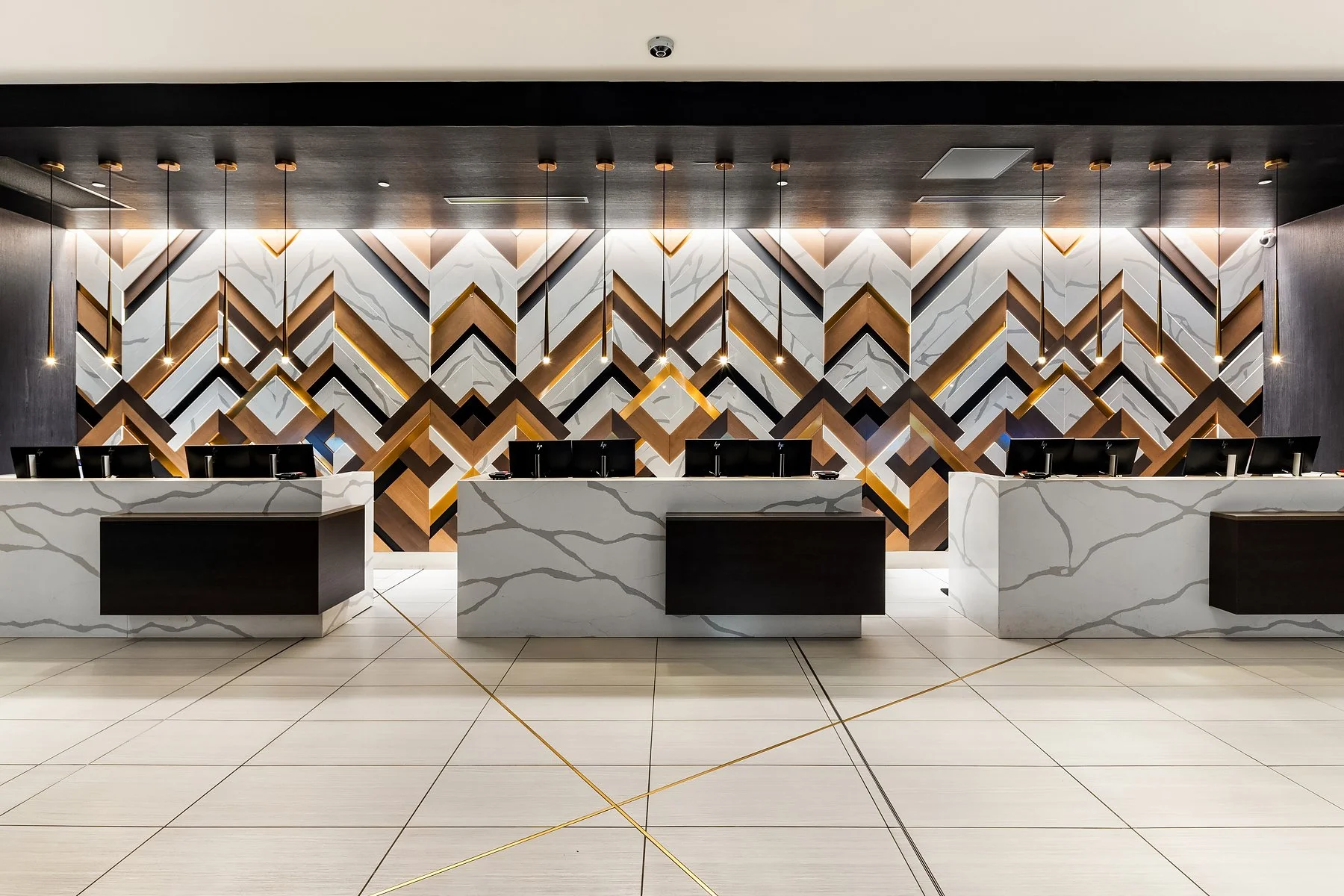 Modern hotel reception with three marble counters and a geometric patterned wall in black, white, and wood tones behind them, illuminated with pendant lights