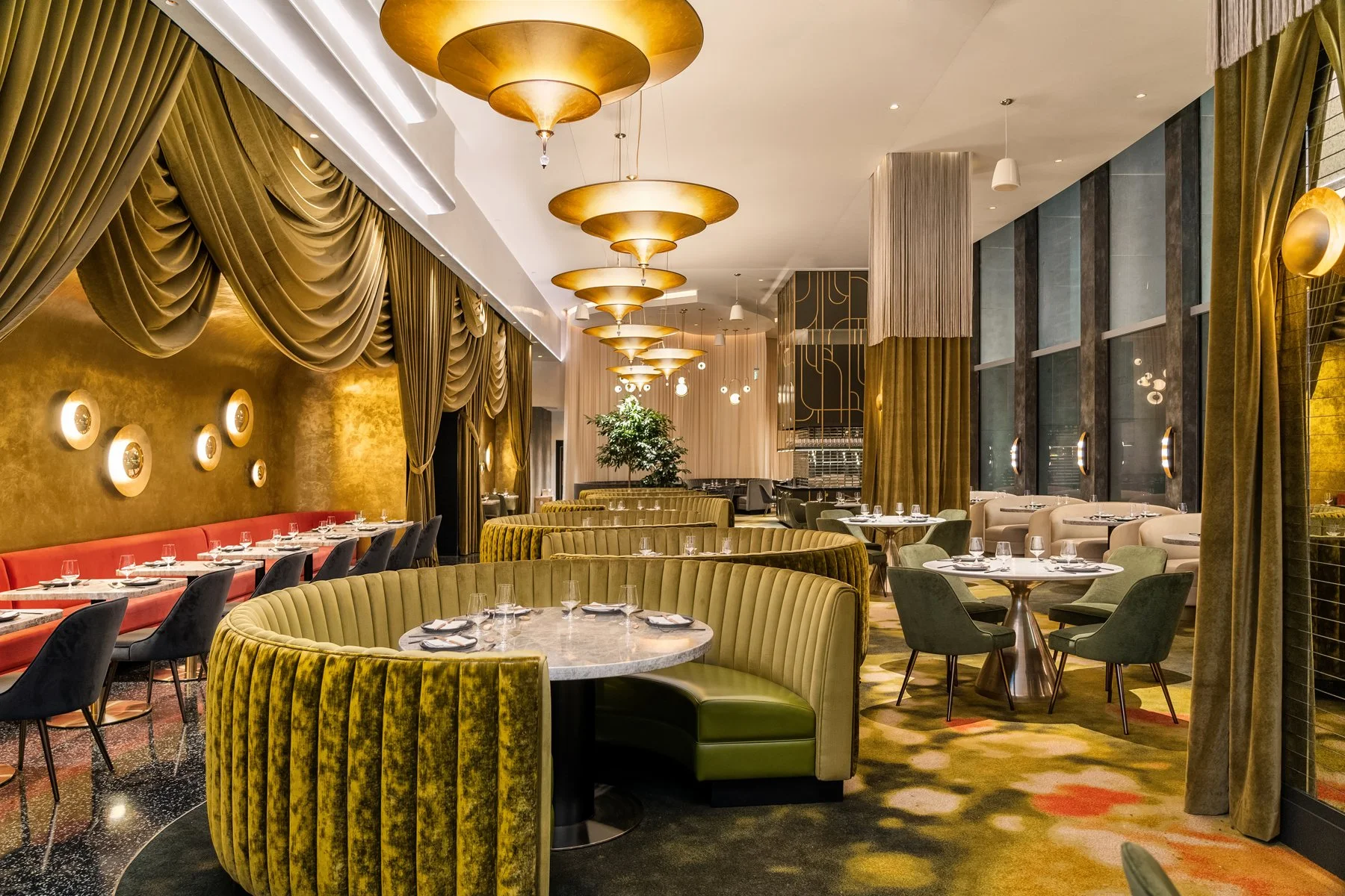 Interior of a modern upscale restaurant with velvet seating in shades of green, red, and beige, round marble tables set with glassware and cutlery, gold-colored ceiling fixtures, gold curtains, and large windows.