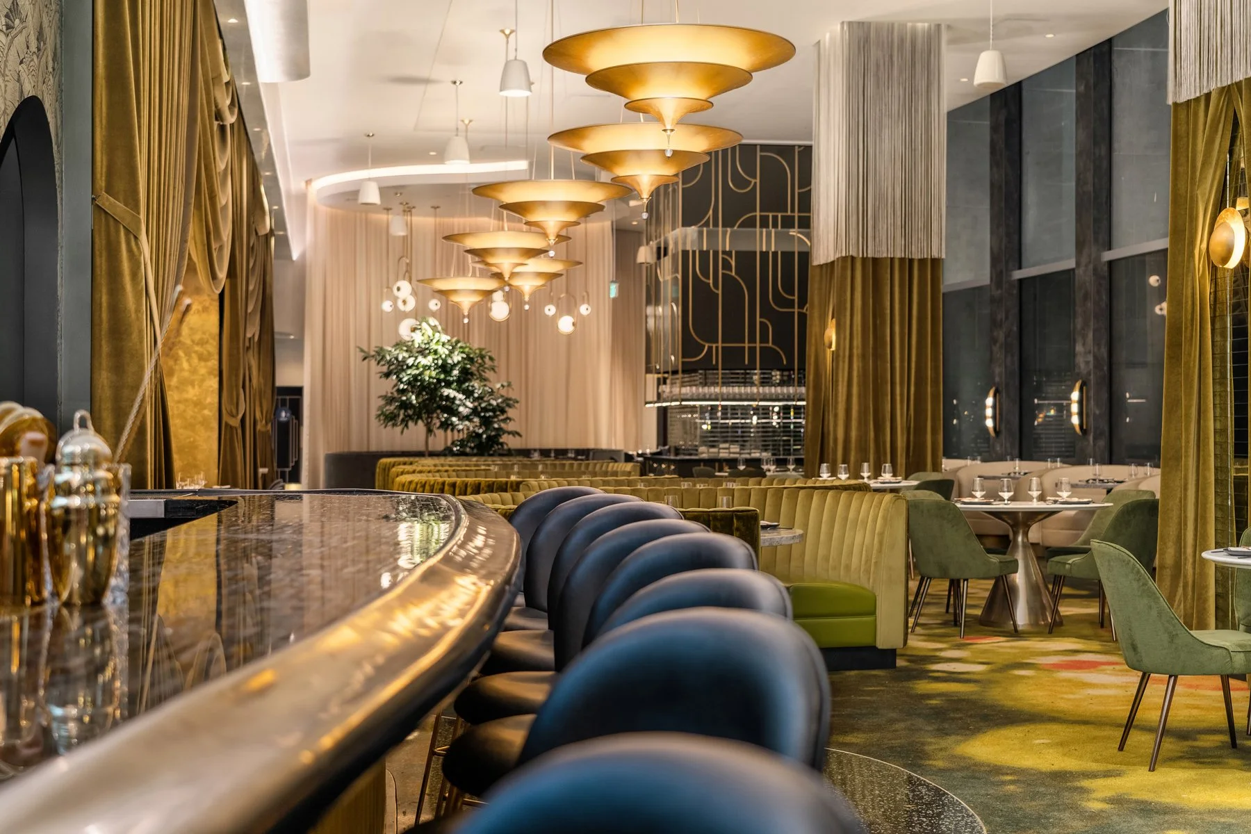 Luxury restaurant interior with a bar counter, black and green velvet chairs, gold accents, and hanging gold light fixtures.