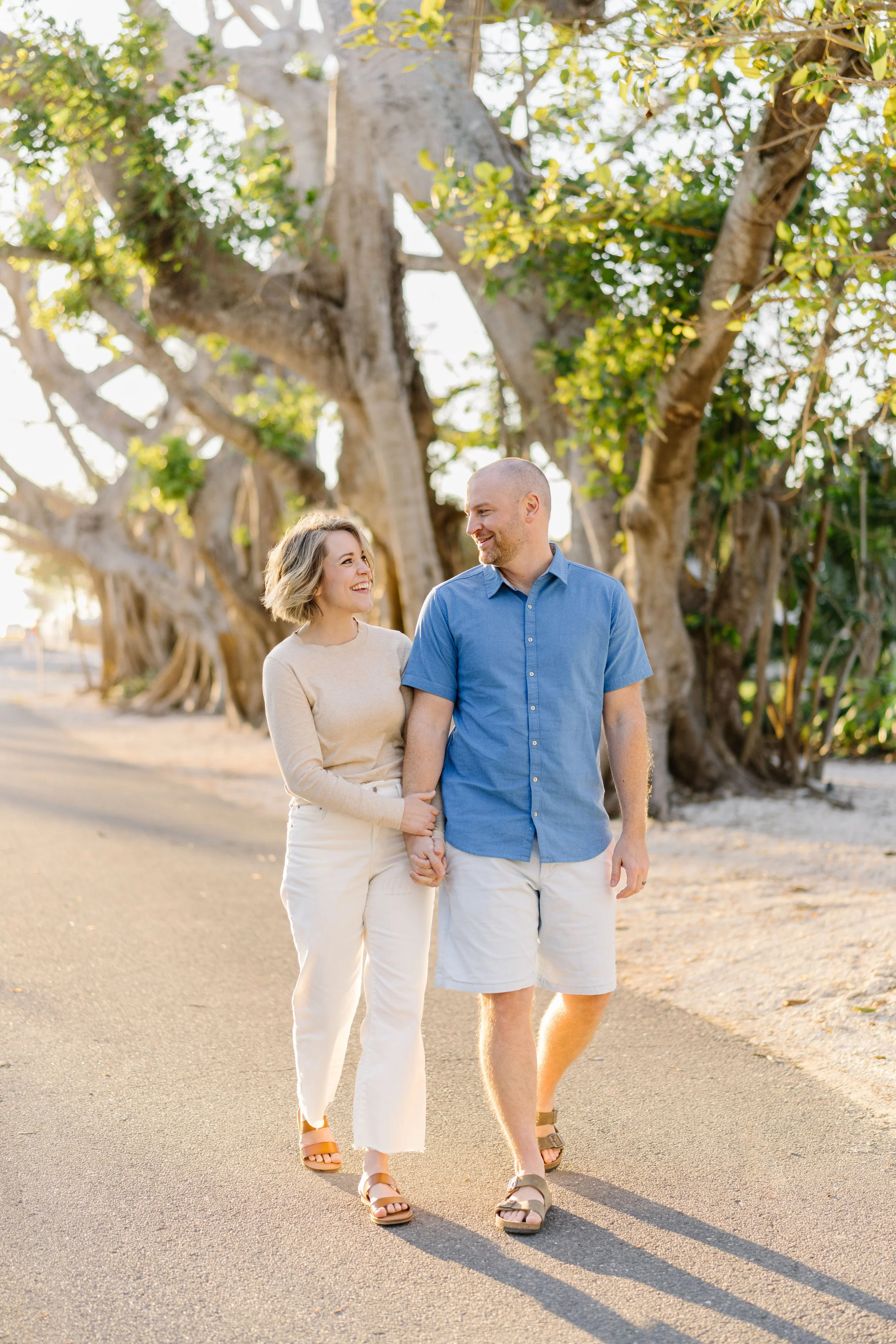 A couple walking along a sidewalk holding hands, smiling, with large trees in the background during sunset.
