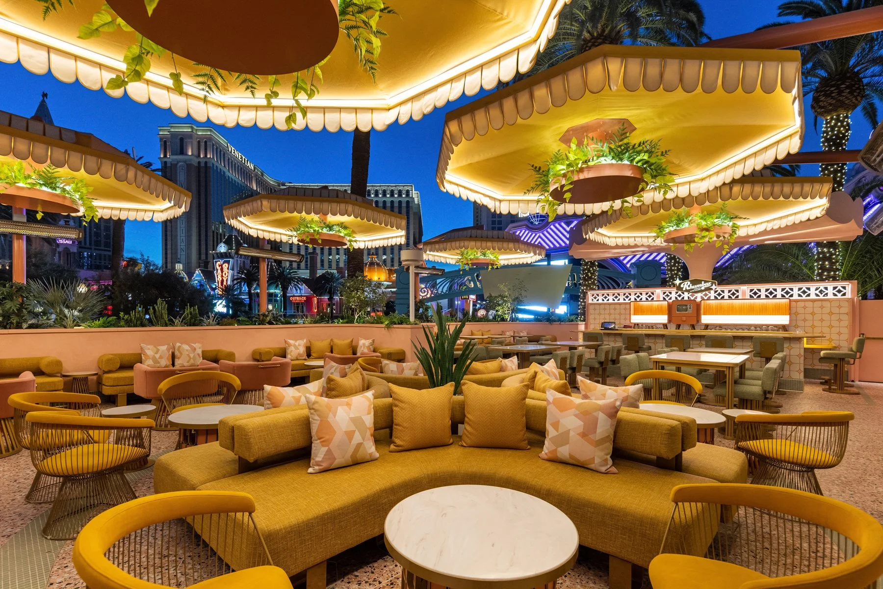 Nighttime view of an outdoor seating area with yellow and green cushioned chairs and sofas, decorated with patterned pillows, under illuminated umbrellas with plants, overlooking city buildings and a lively street.
