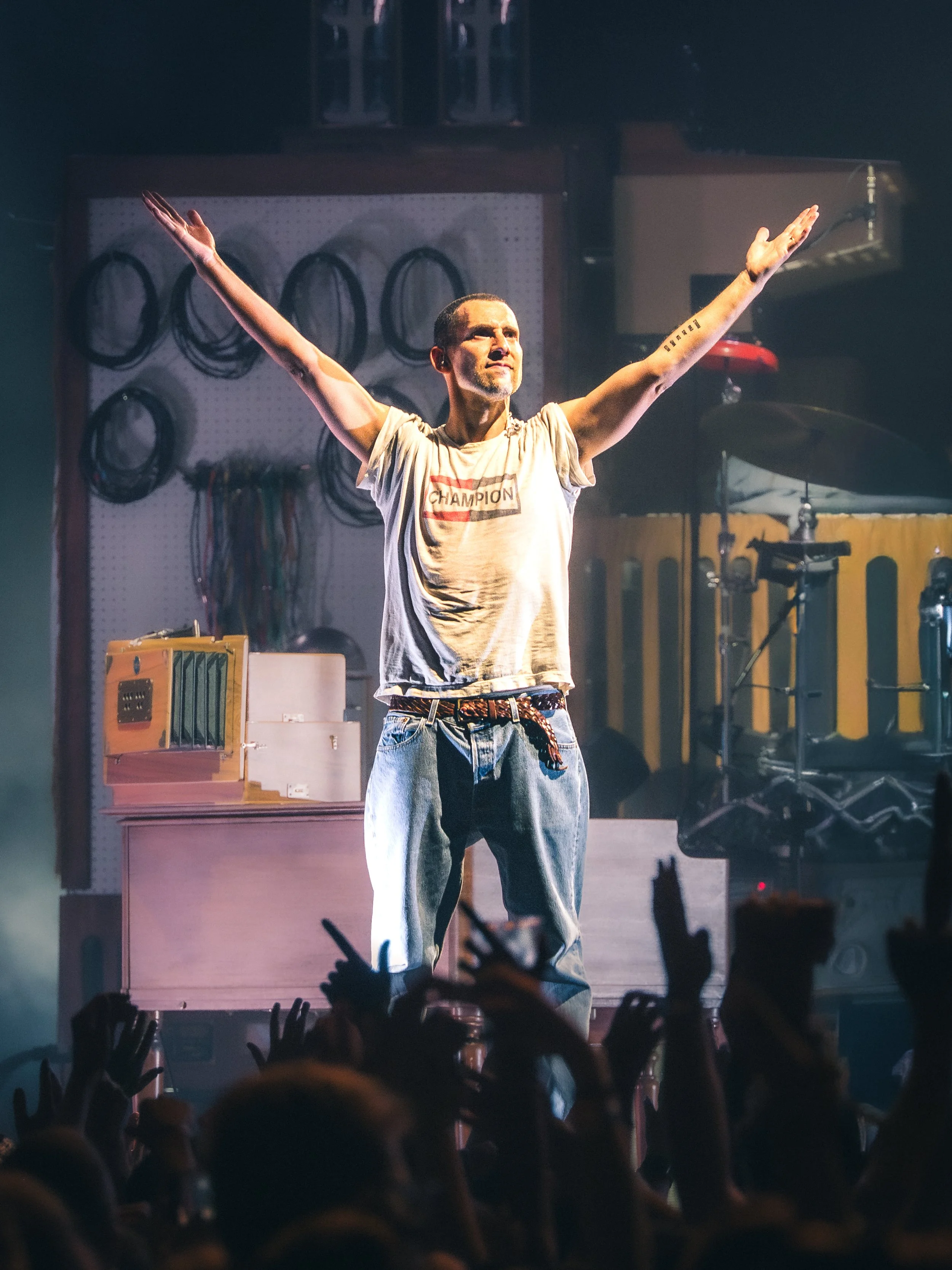 A man with short hair and a beard stands on a stage with his arms raised, in front of an audience. He wears a gray T-shirt with a red and black logo and light blue jeans. The background includes musical equipment and a wall with circular wire hangers.