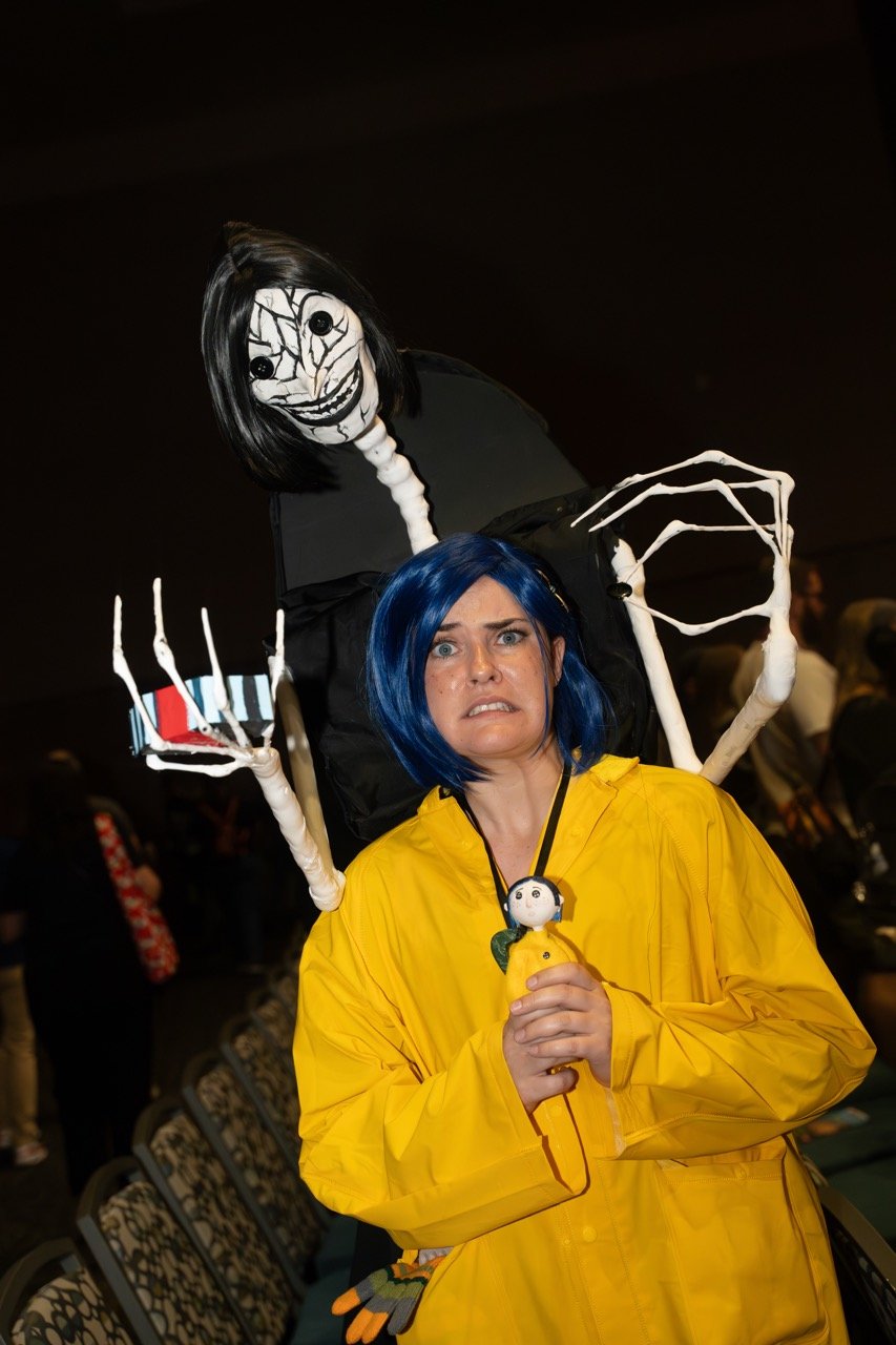 A woman dressed as a character from the movie 'Coraline' with blue hair and a yellow raincoat, holding a small doll of Coraline, looking frightened. Behind her is a large puppet of the Other Mother, a creepy figure with black hair and a face with cracks, holding out claws.