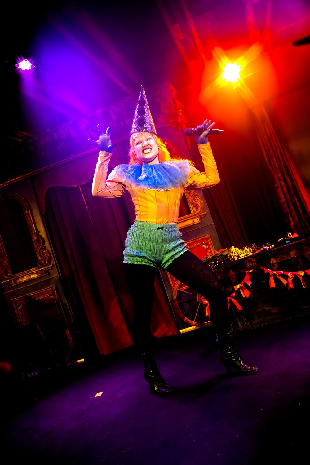 A performer dressed as a clown with a pointy hat, yellow top, blue ruffled collar, green shorts, and black tights dancing on stage with colorful lighting.