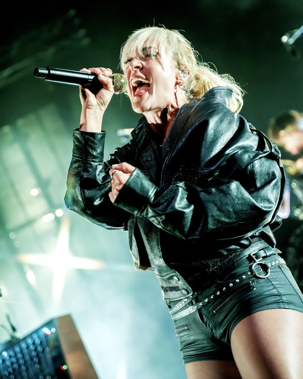 Female singer performing passionately on stage, wearing a black leather jacket and shorts, holding microphone, with stage lighting in the background.