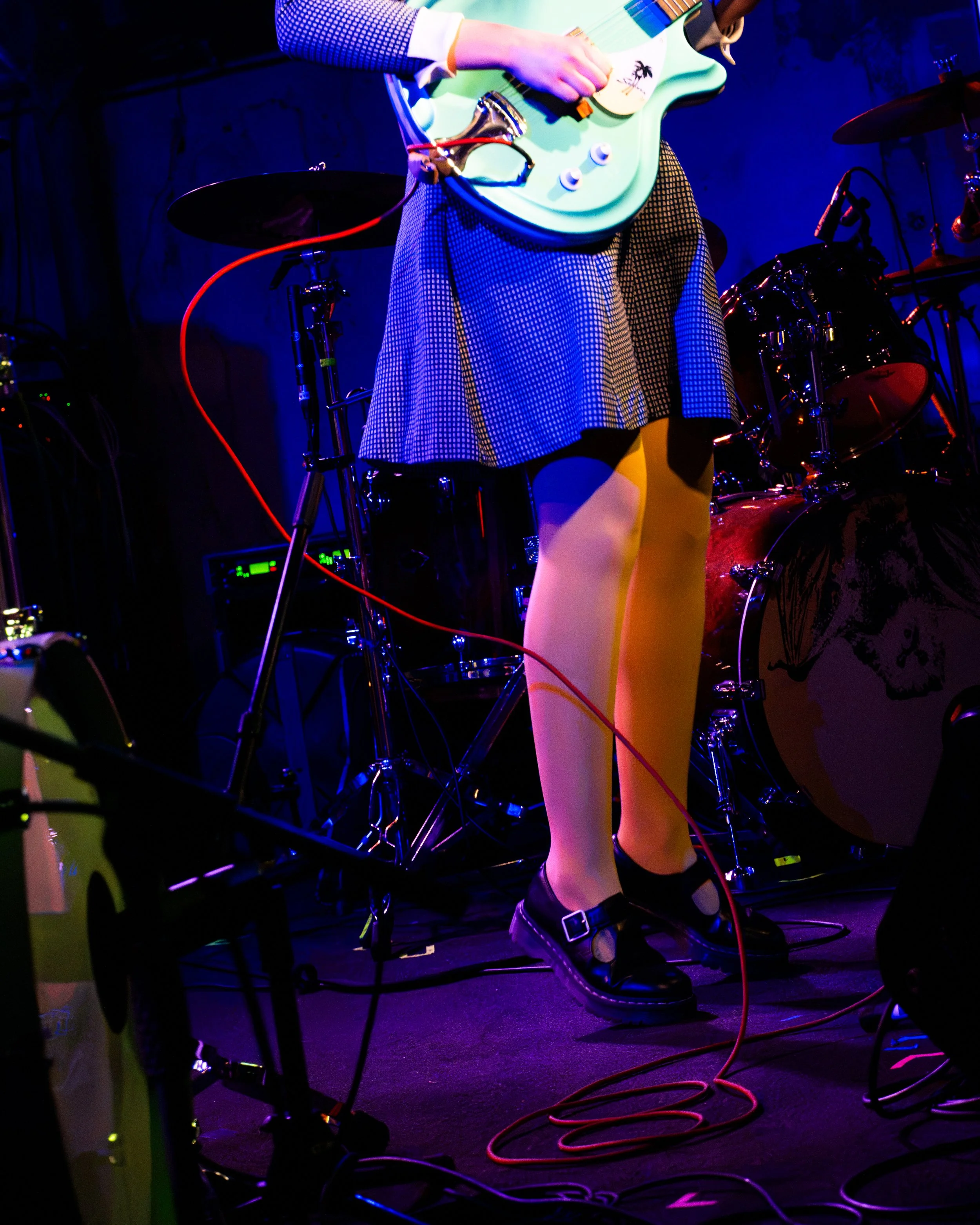 A woman playing an electric guitar on stage during a live music performance. She is wearing a checkered skirt, yellow tights, and black shoes, under colorful stage lighting. Cables and musical equipment are visible around her.