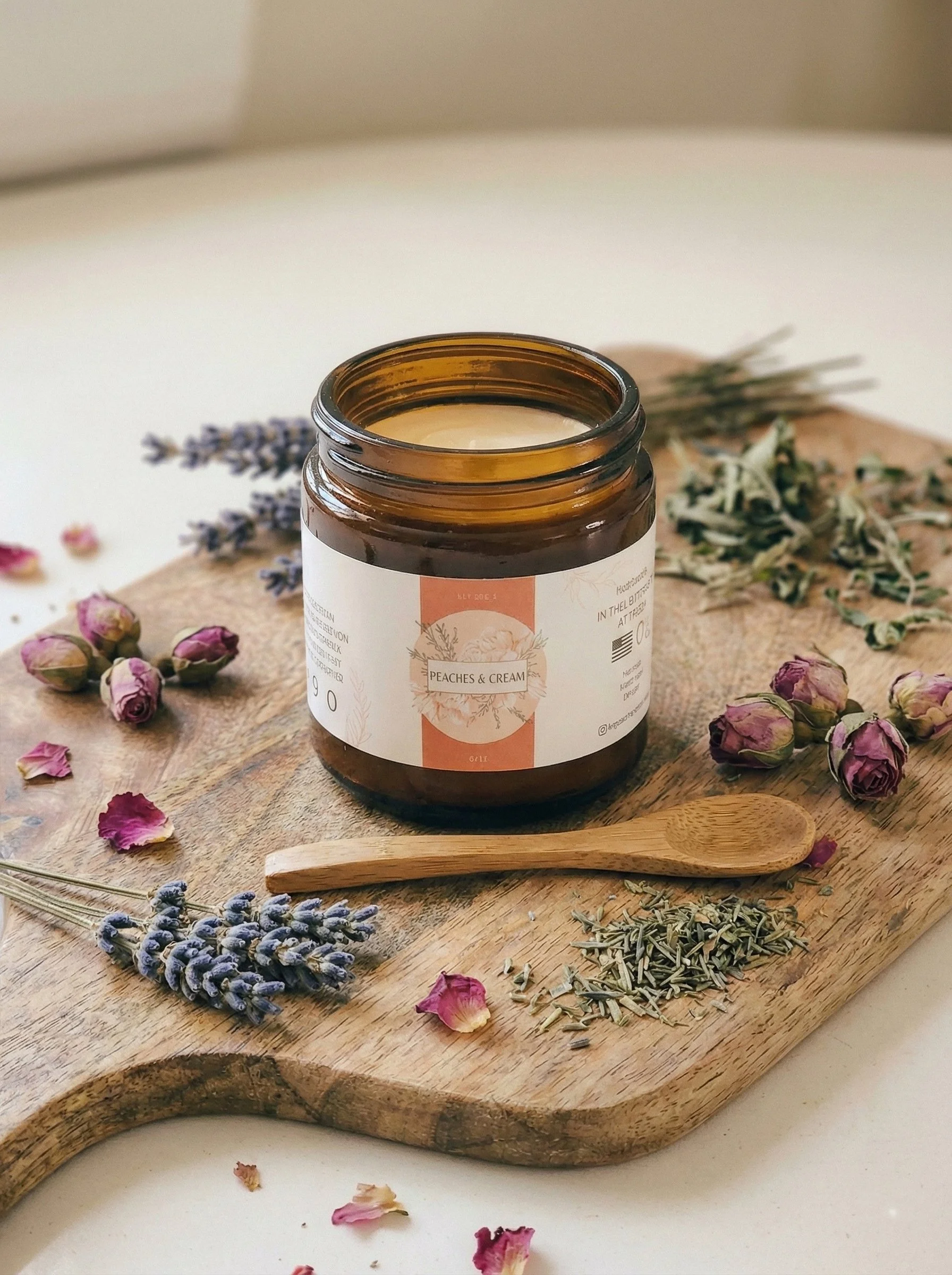 A jar labeled 'Peaches & Cream' candle on a wooden board, surrounded by dried lavender, pink rosebuds, and dried herbs, with a small wooden spoon resting on the board.