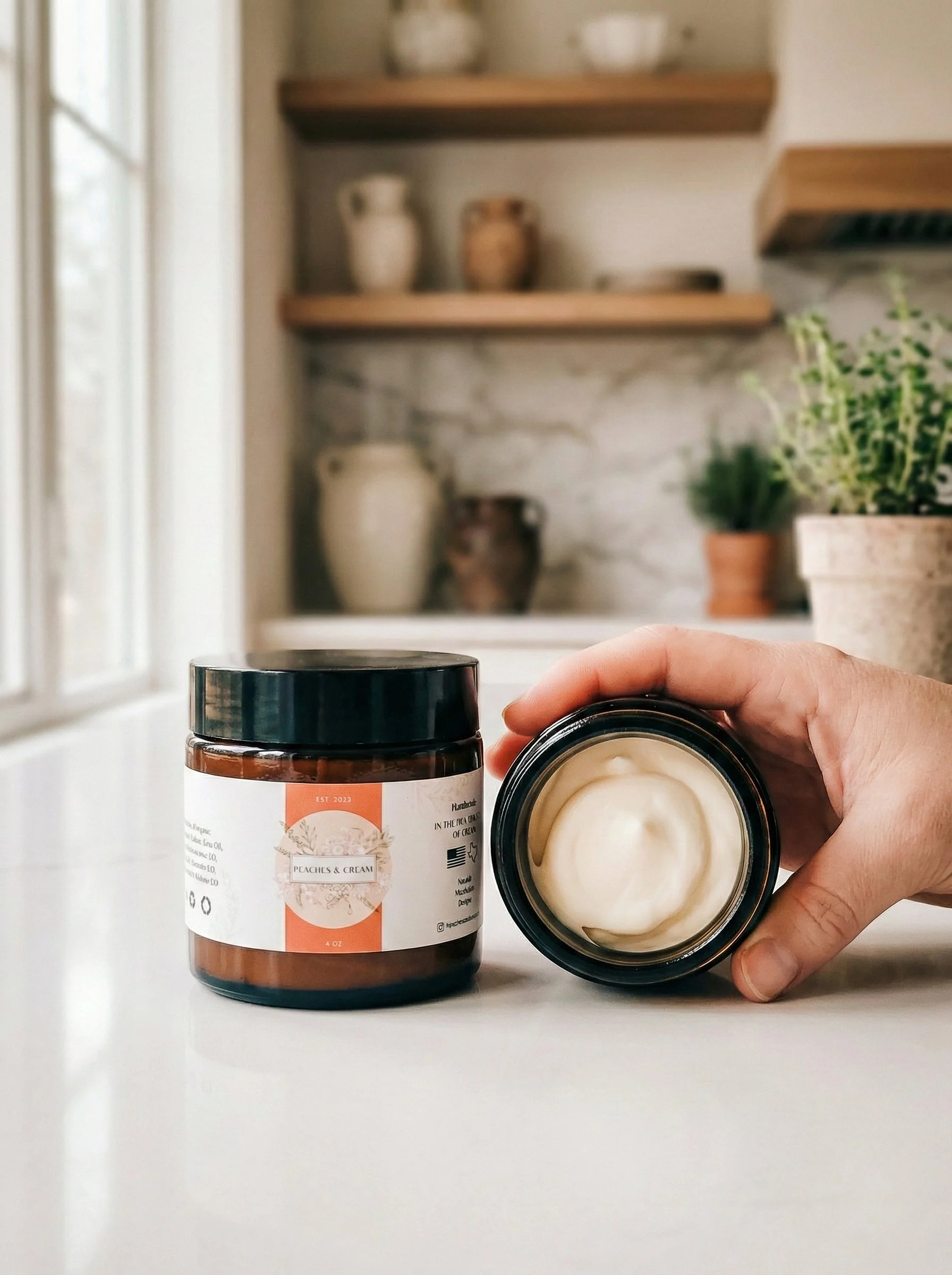 A person's hand holding an open jar of cream next to a closed jar labeled 'Peaches & Cream' on a white countertop in a kitchen with shelves, potted plants, and ceramic jars in the background. Skincare. Organic. Local. Shop. Clean. Essential Oils. Han