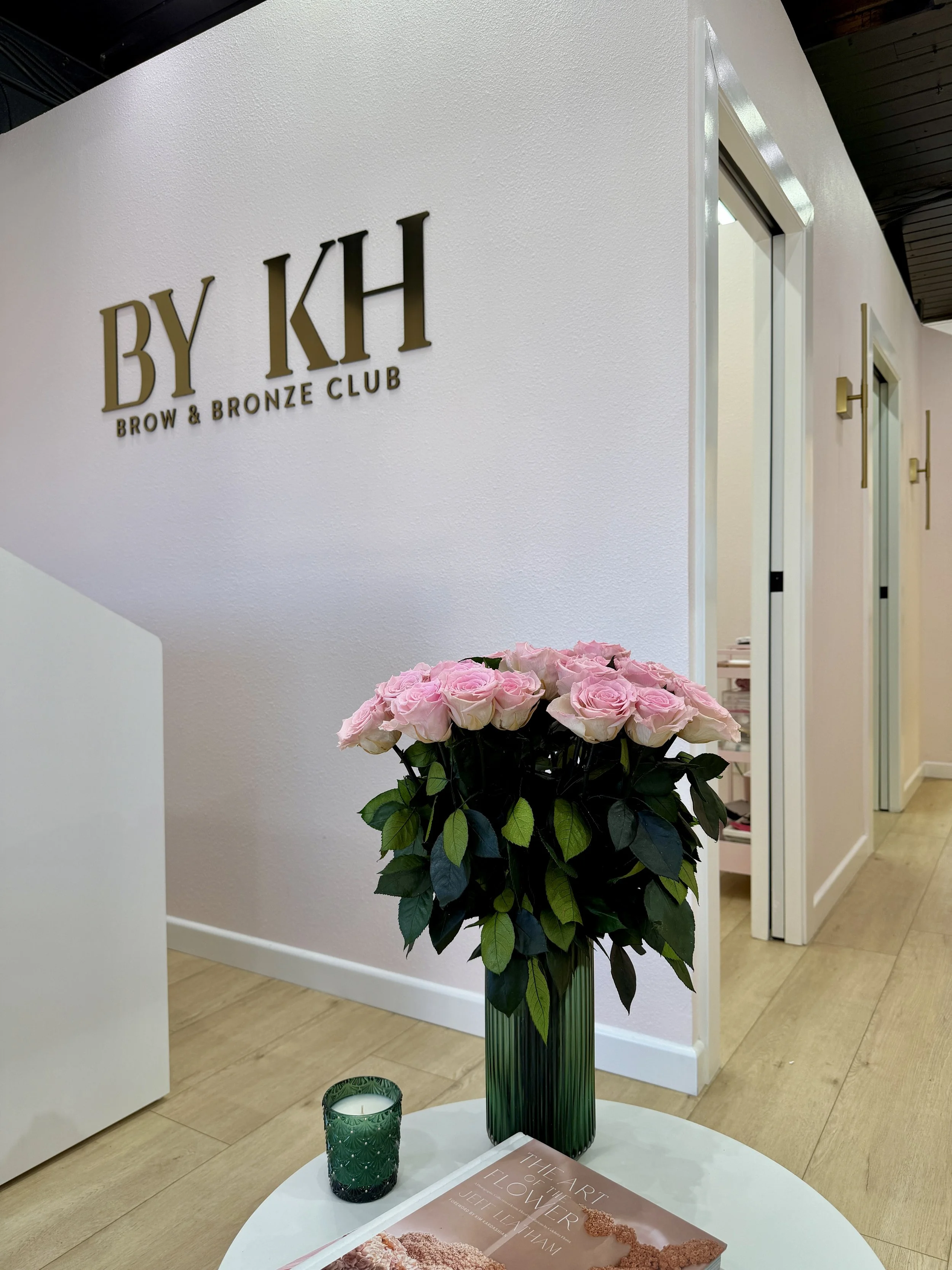 Interior of a beauty salon called BY KH Brow & Bronze Club, with pink roses in a green vase, a green candle holder, and a magazine on a white table.