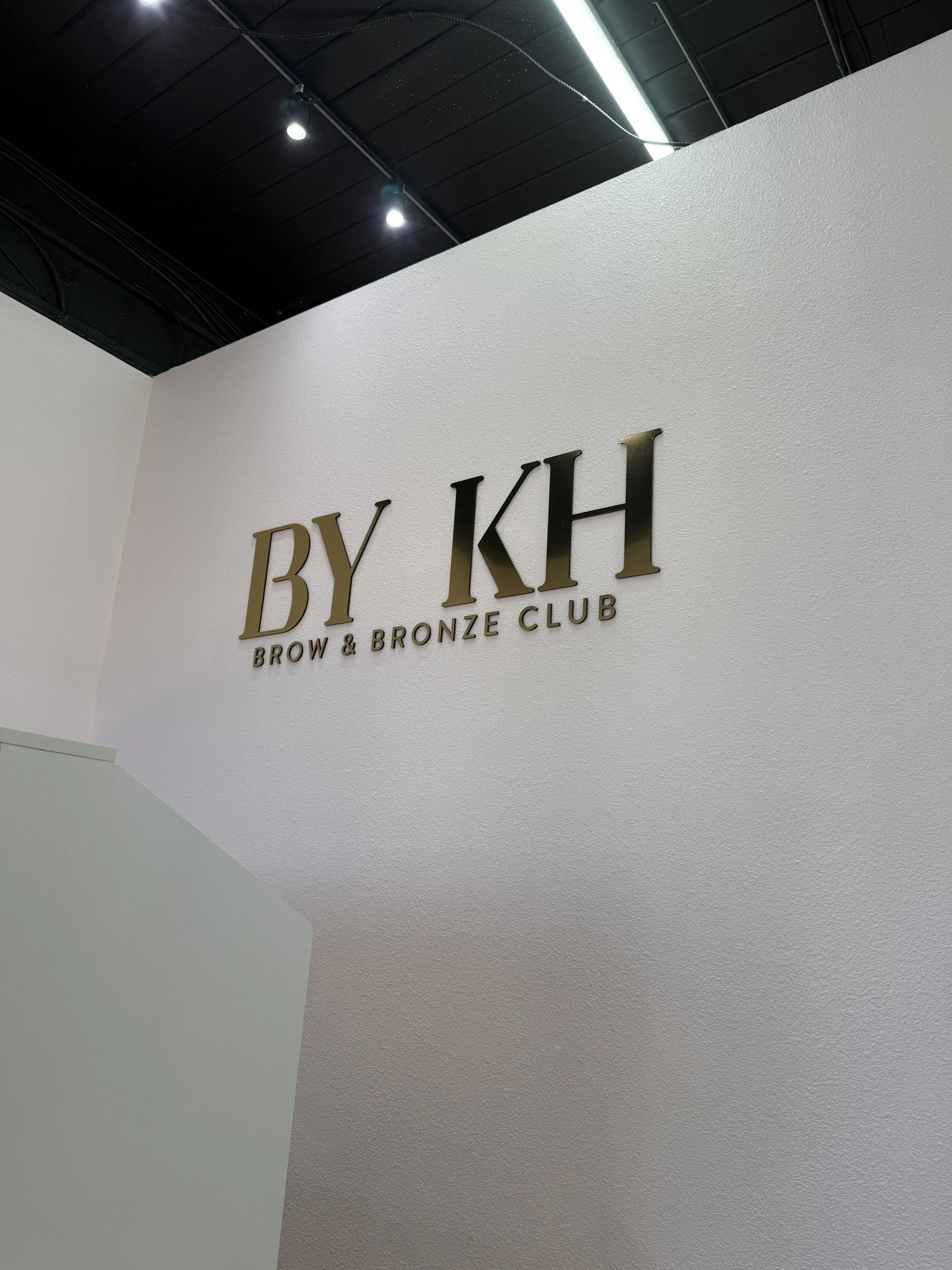 Interior of a salon or club with a wall sign reading 'BY KH Brow & Bronze Club' and a black ceiling with lighting.