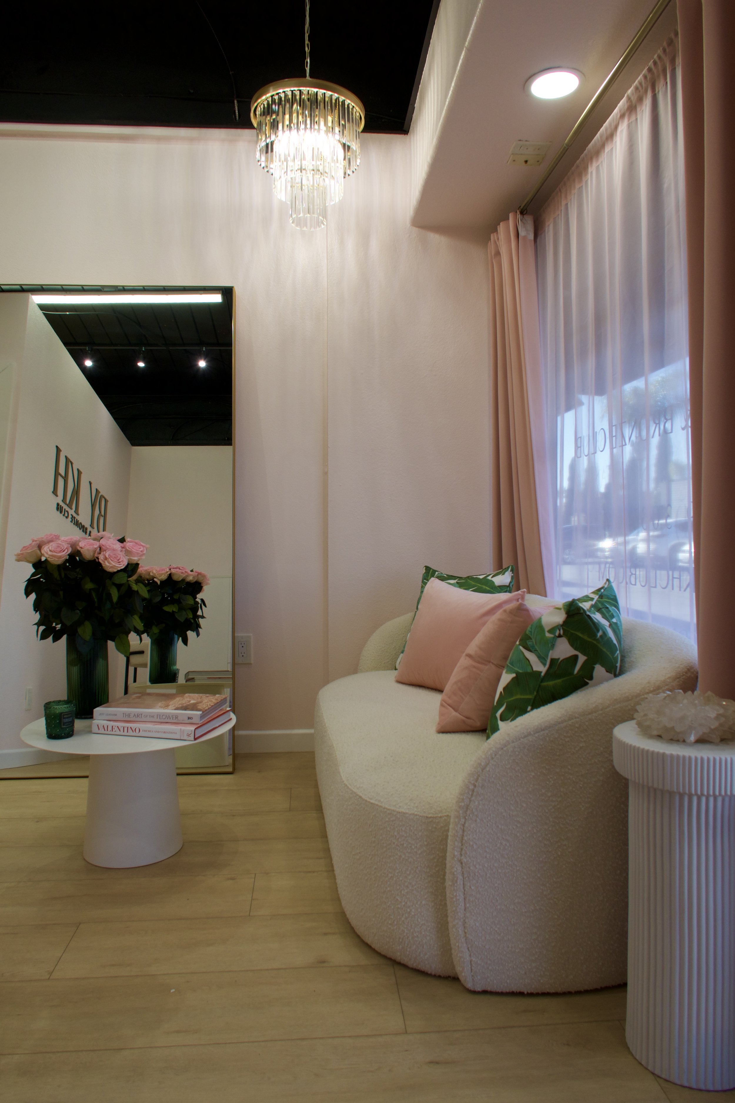 A cozy interior space with a cream-colored curved sofa decorated with pink and green leaf-patterned pillows, a side table with a seashell ornament, a bouquet of pink roses, and a large mirror reflecting the room. Pink curtains hang by a window, and a