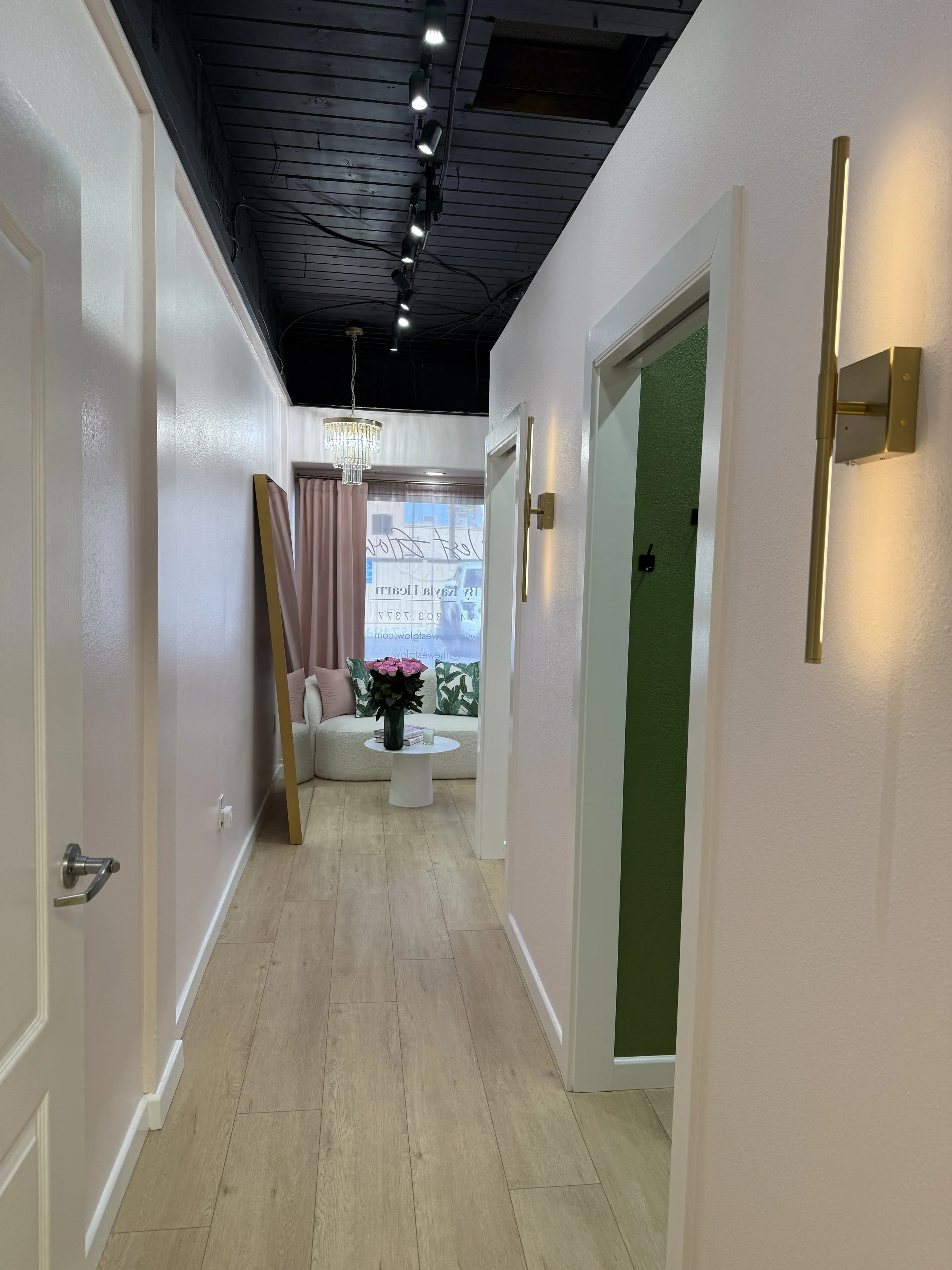 Hallway leading to a sitting area with a white sofa, pink and green pillows, a street view through a window, and pink curtains. Decor includes a chandelier, wall sconces, and a large mirror leaning against the wall.