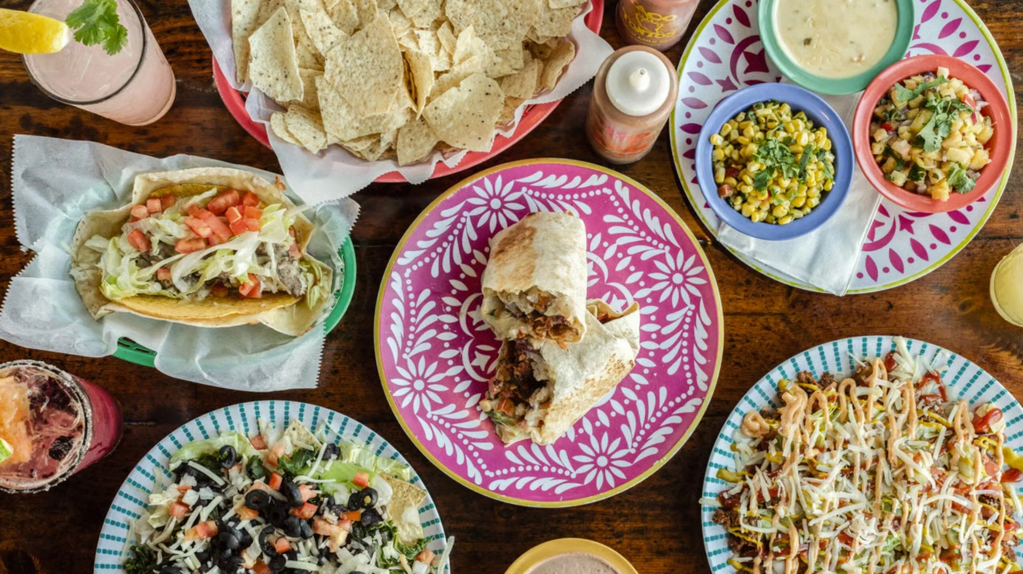 A table full of delicious Taco Tontos food, including chips and dip, a BLT burrito, a crunchy taco with steak, a Cheeseburgerito bowl, house salsa, and refreshing cocktails